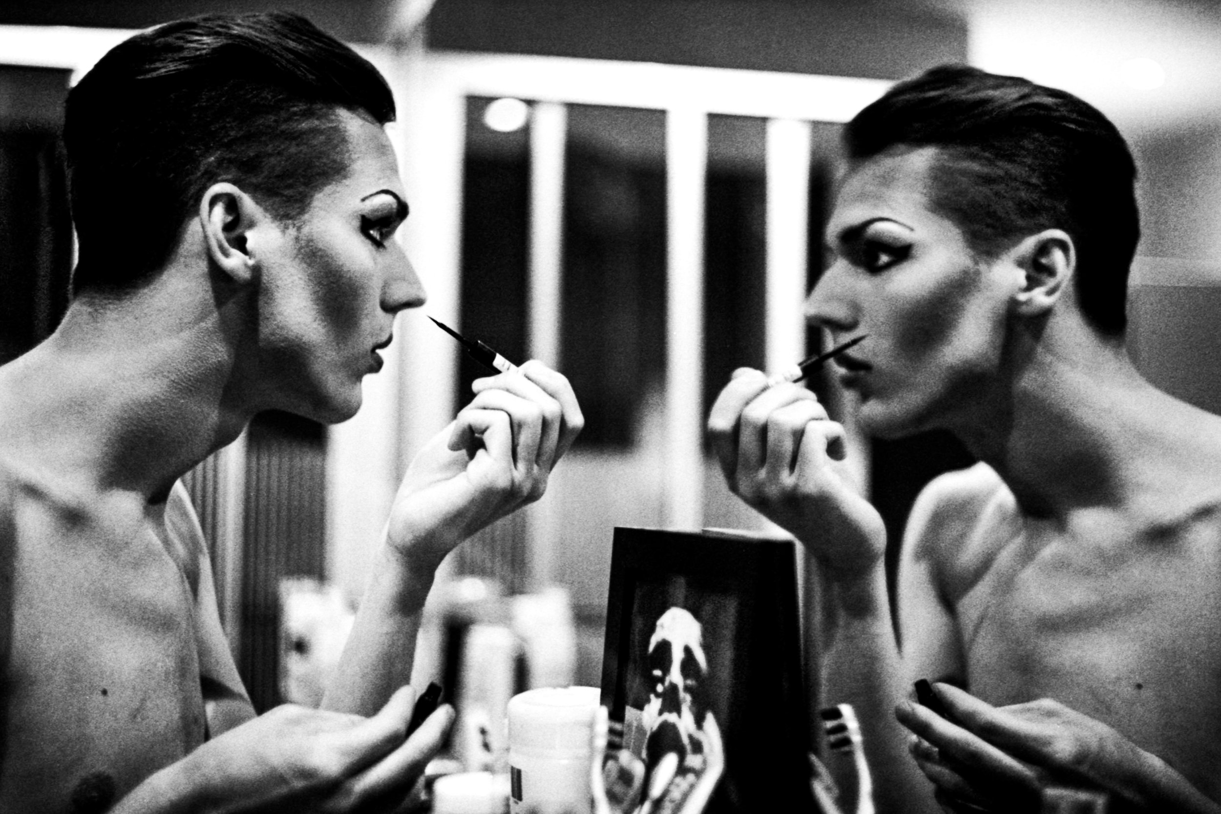 Two women with similar hairstyles and makeup, holding small makeup brushes close to their lips, looking into each other's eyes in a black and white photo.