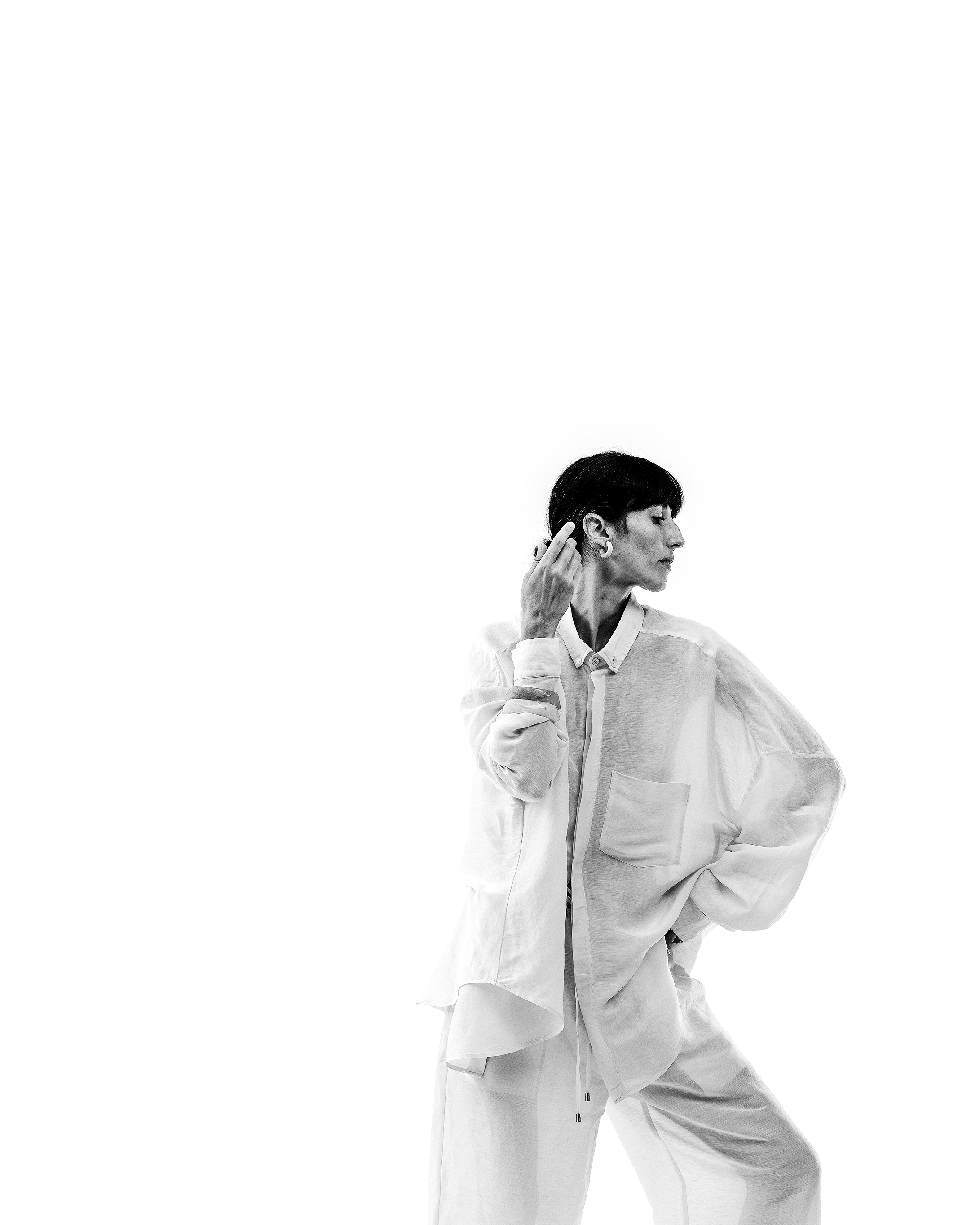 Black and white photo of a woman with short dark hair wearing a loose white shirt and pants, standing against a plain white background, looking to her right, with one hand touching her ear and the other on her hip.