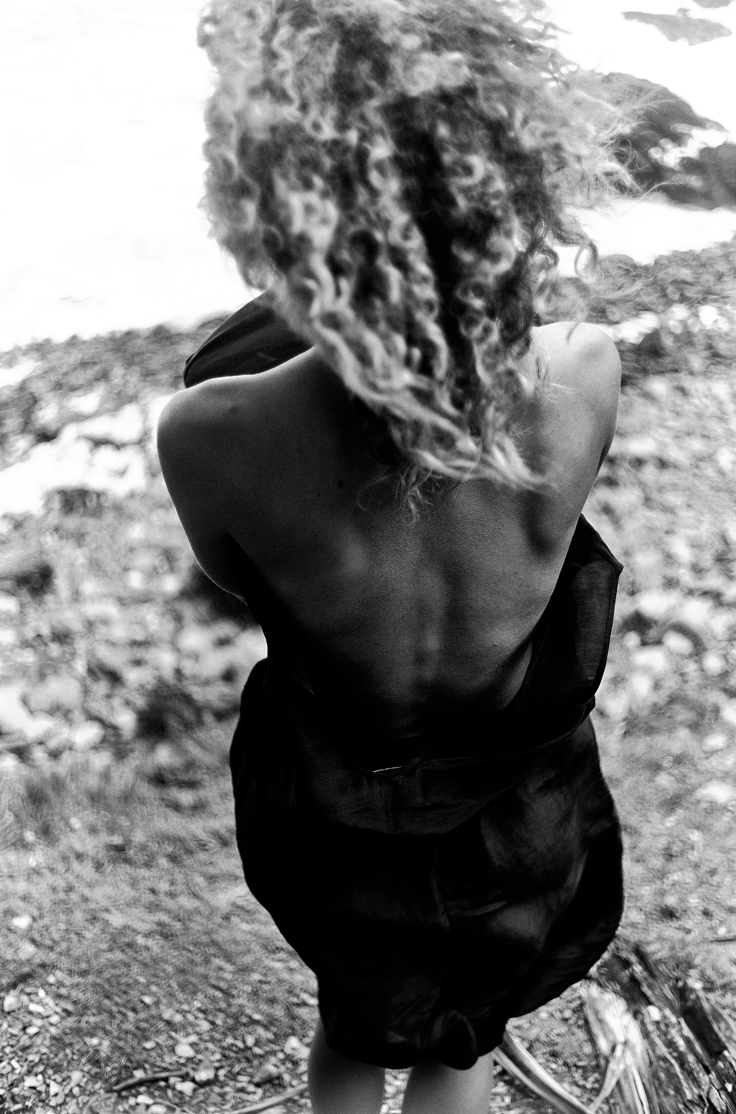 A person with curly hair and an arm tattoo is crouching outdoors on a dirt and gravel surface, facing away from the camera.