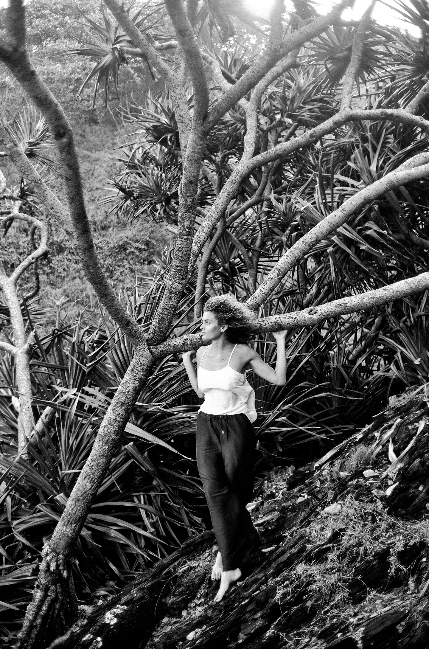 A woman with curly hair wearing a sleeveless top and loose pants, standing barefoot in a tropical forest with dense foliage and tree branches around her.