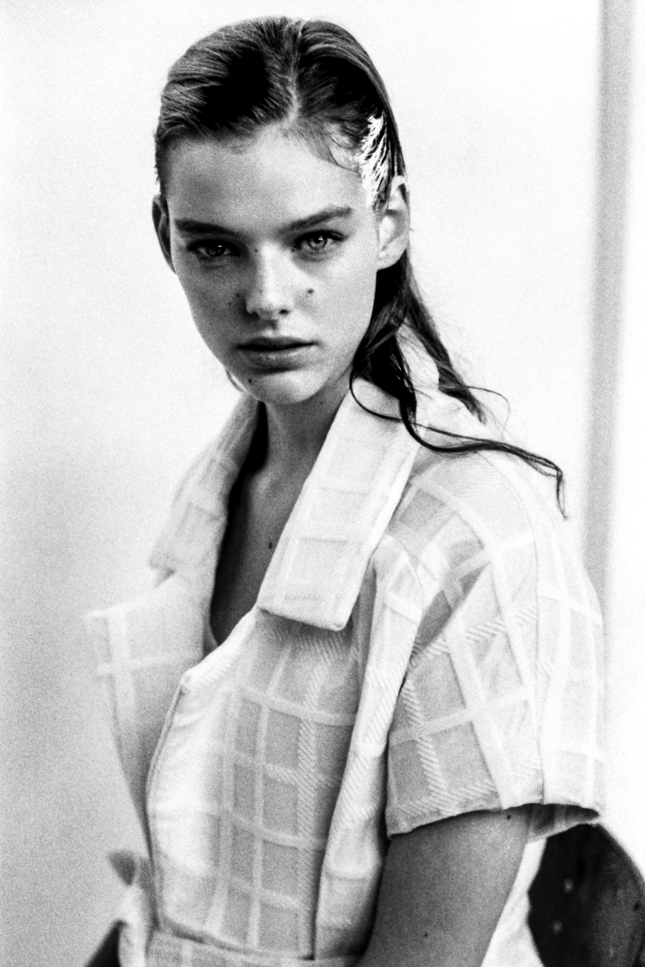 A black-and-white portrait of a young woman with sleek, wet-looking hair, wearing a checked blazer over a dark top, looking directly at the camera with an intense expression.