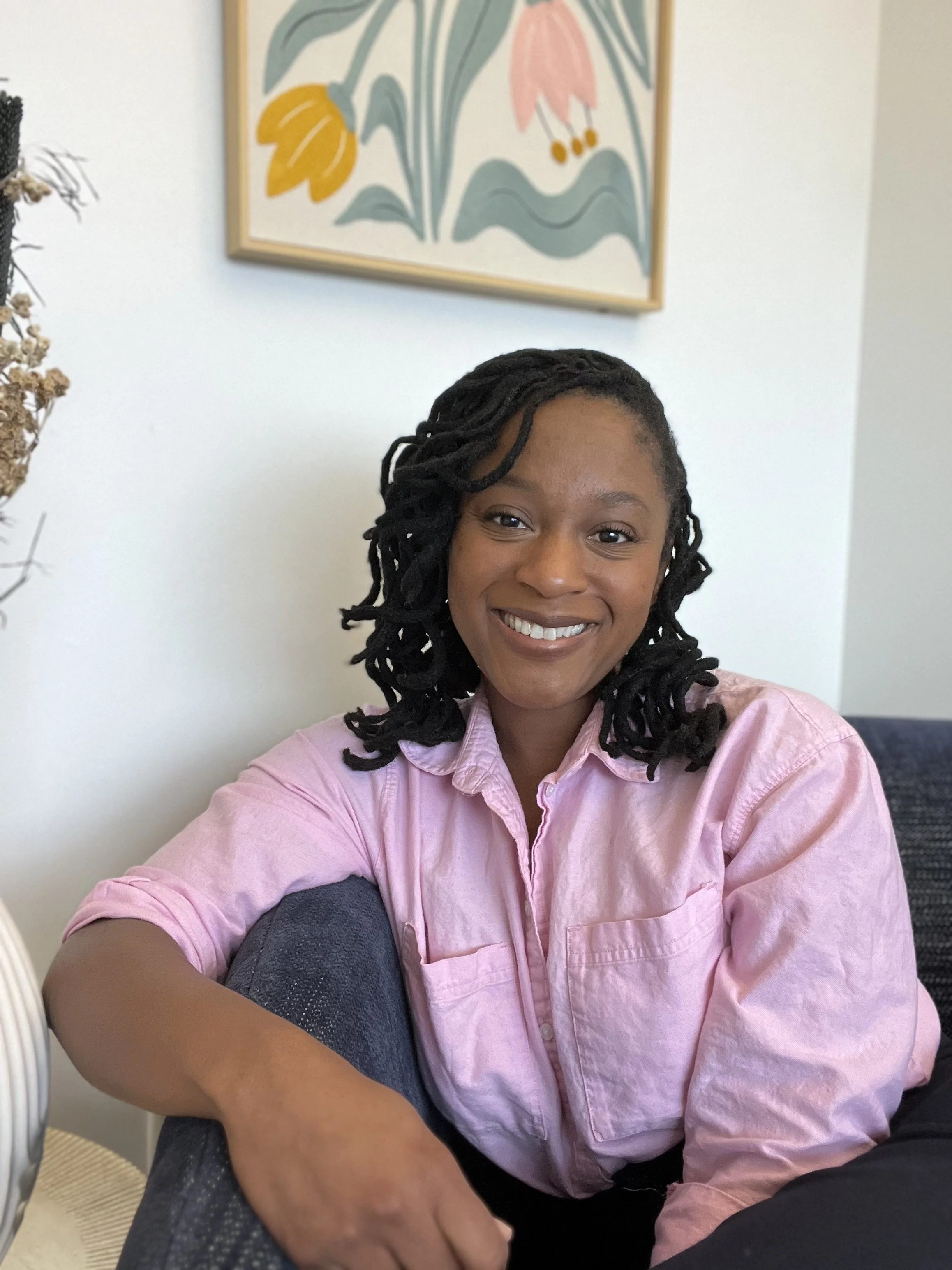 A Black Expressive Arts Therapist located in California gently smiling at the camera while seated in a blue chair