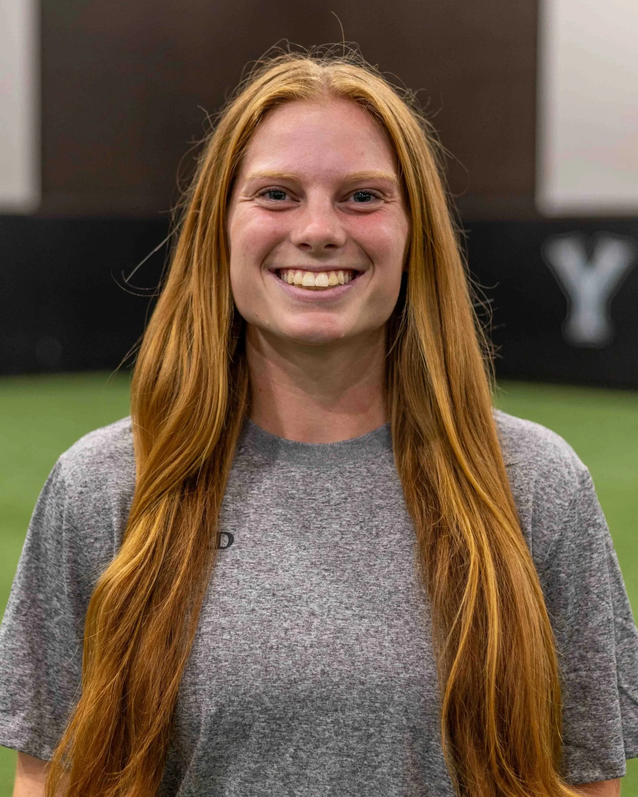 A young woman with long red hair, smiling, standing in an indoor sports facility. softball player at uga. middle infielder. softball instructor for hitting and infield defense
