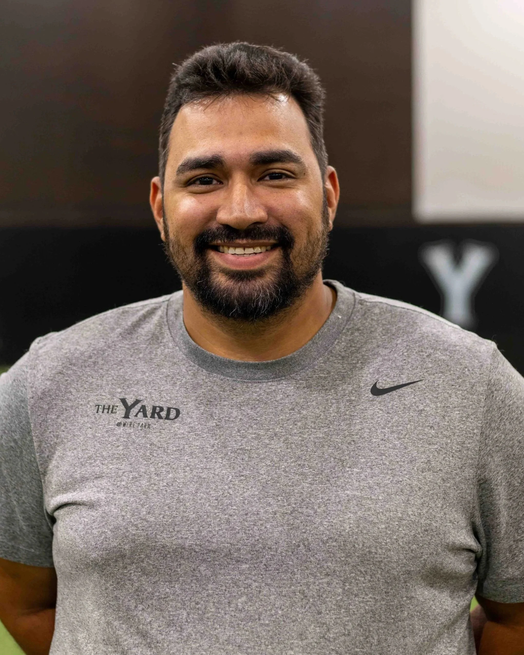 A man with a beard smiling, wearing a gray Nike t-shirt with 'The Yard', baseball instructor. softball instructor. hitting instructor. fielding instructor