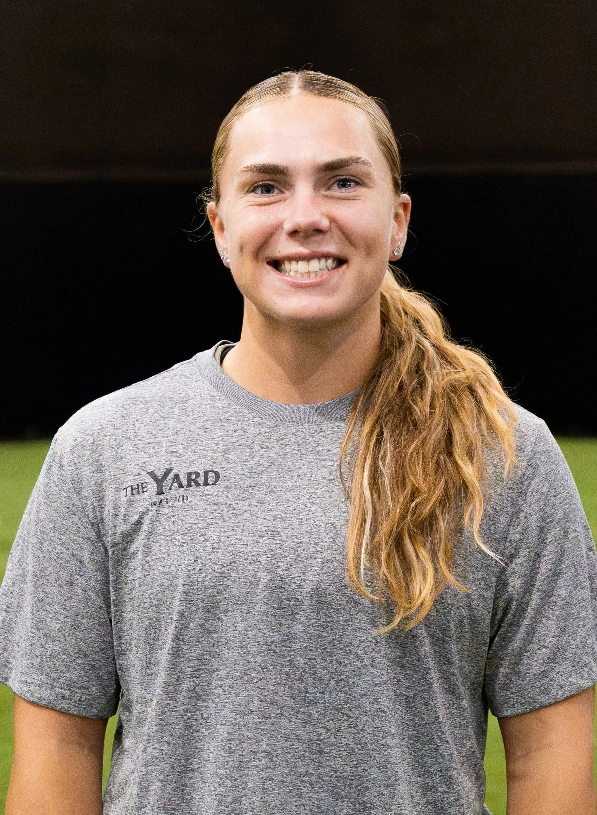A young woman with long, wavy, blonde hair tied in a side ponytail, smiling and wearing a gray T-shirt with 'The Yard' logo, standing on a grassy field at night. current uga softball player and instructor