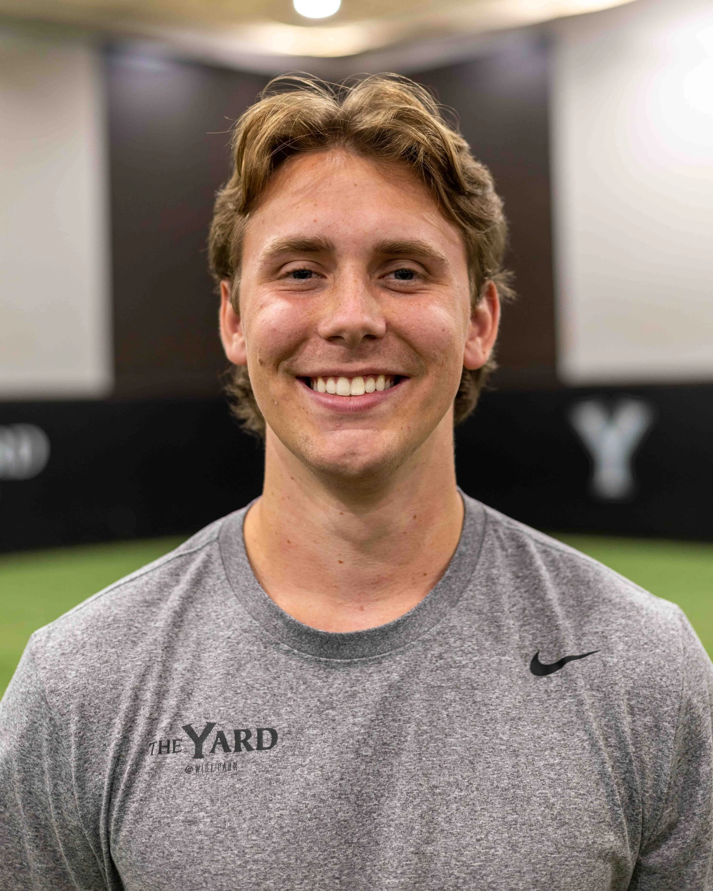 Young man with light brown hair and a gray athletic shirt smiling in an indoor baseball facility. max puckett. baseball facility.