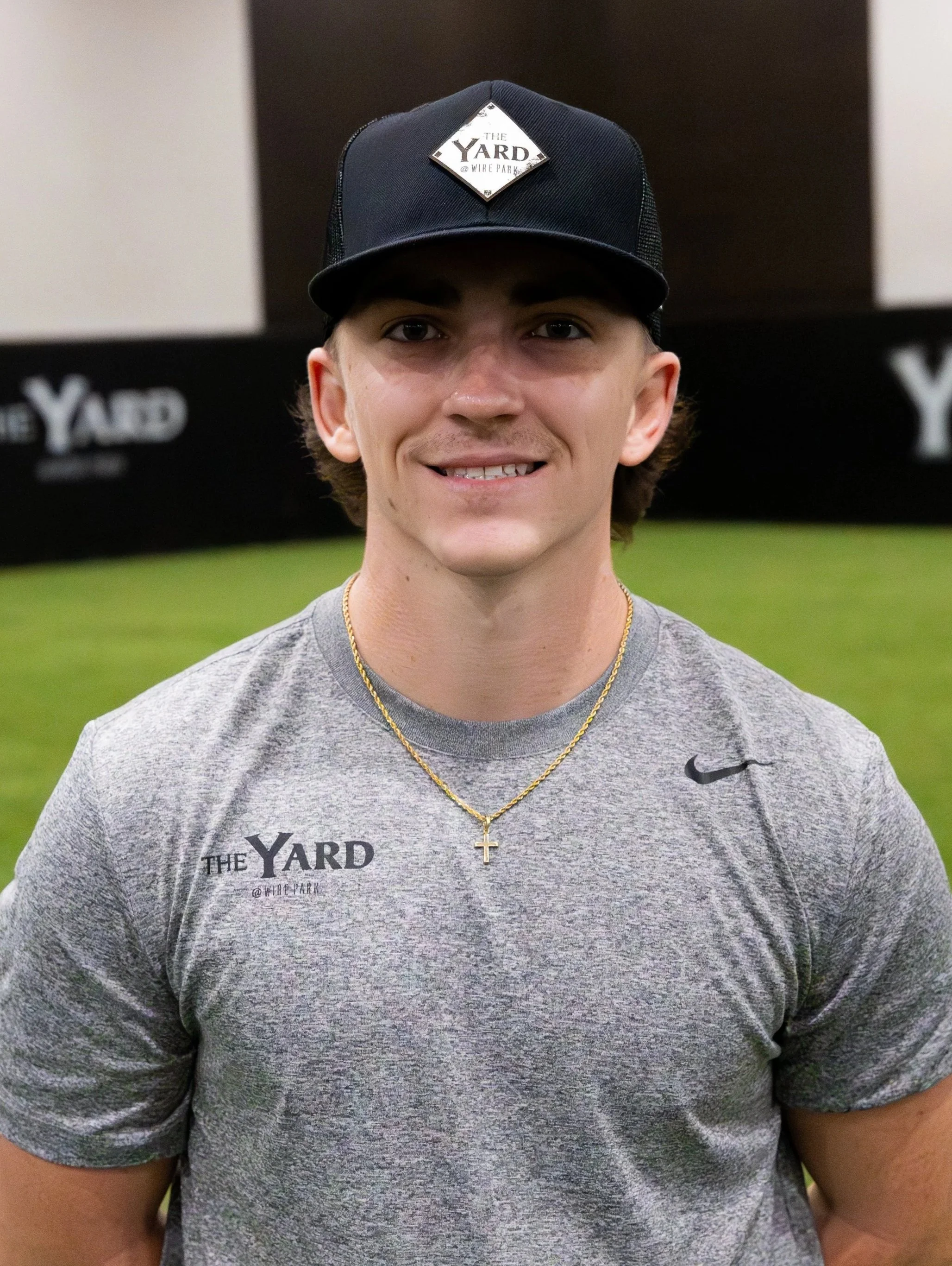 A young man wearing a black baseball cap with a logo, a gray athletic t-shirt, a gold chain necklace with a cross, smiling, black wall that says 'The Yard' in the background. kyle stephenson, baseball pitching instructor