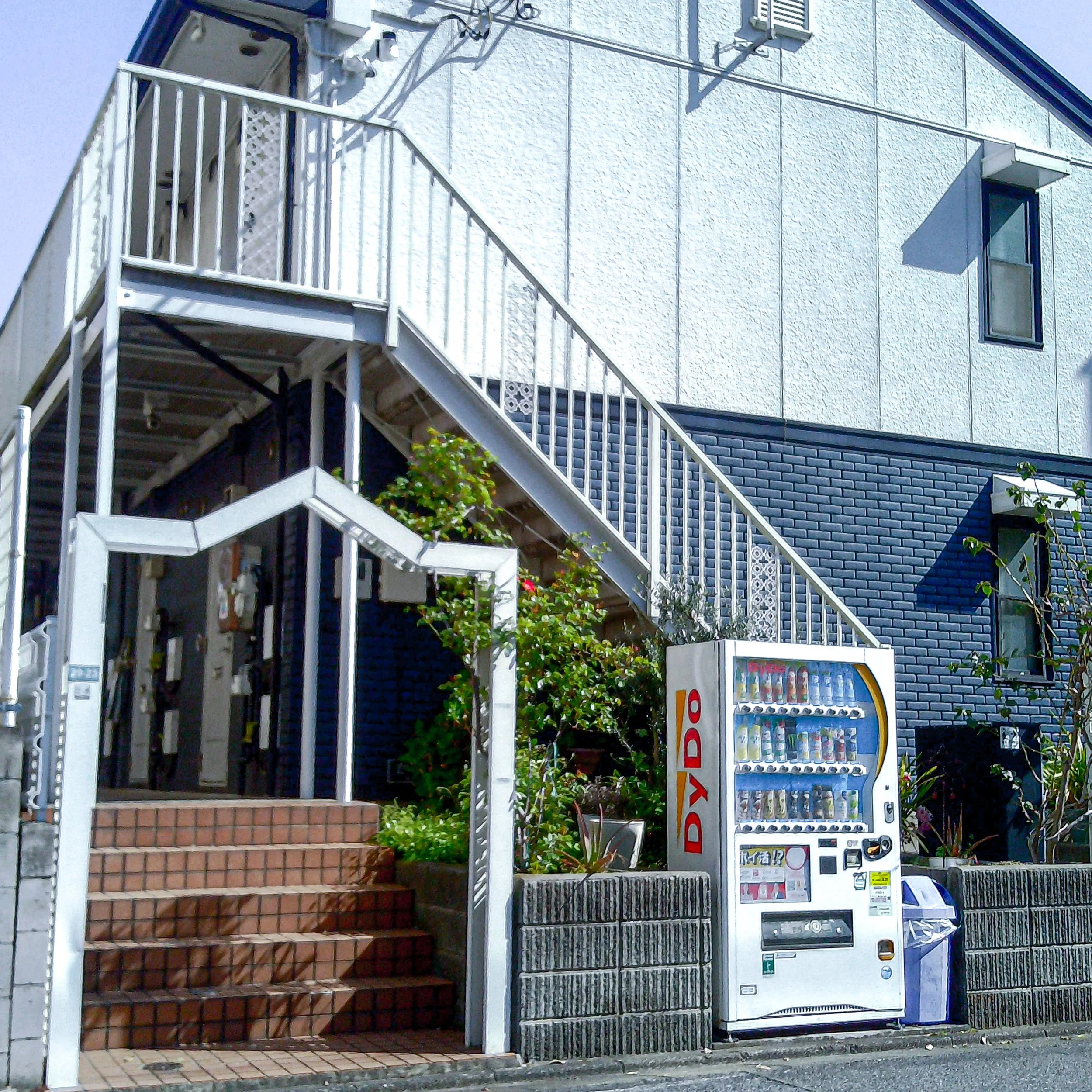 A vending machine outside a residential building with stairs, plants, and a small window.