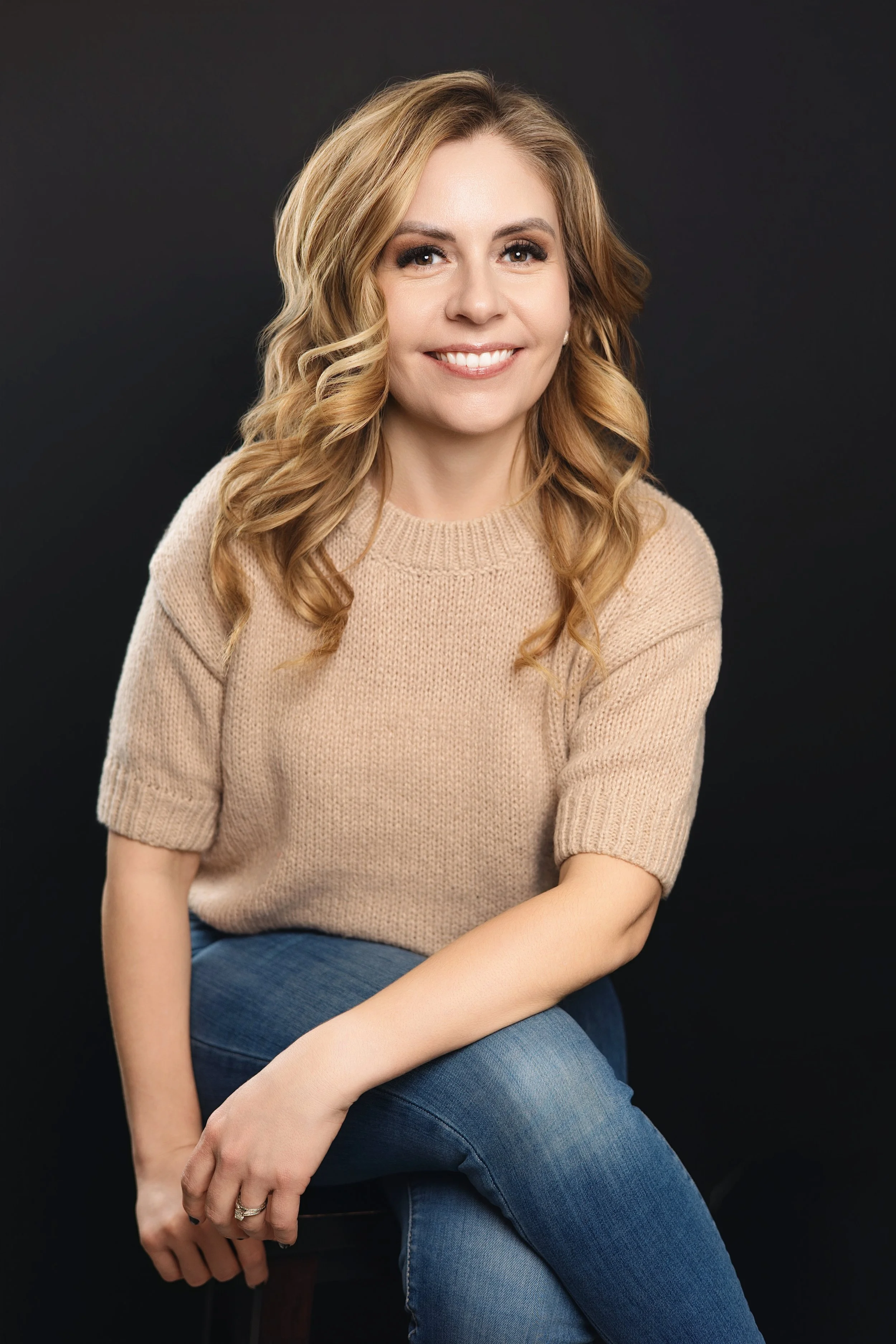Woman with blonde hair and bangs, wearing a dark top and green pants, sitting on a stool, smiling.