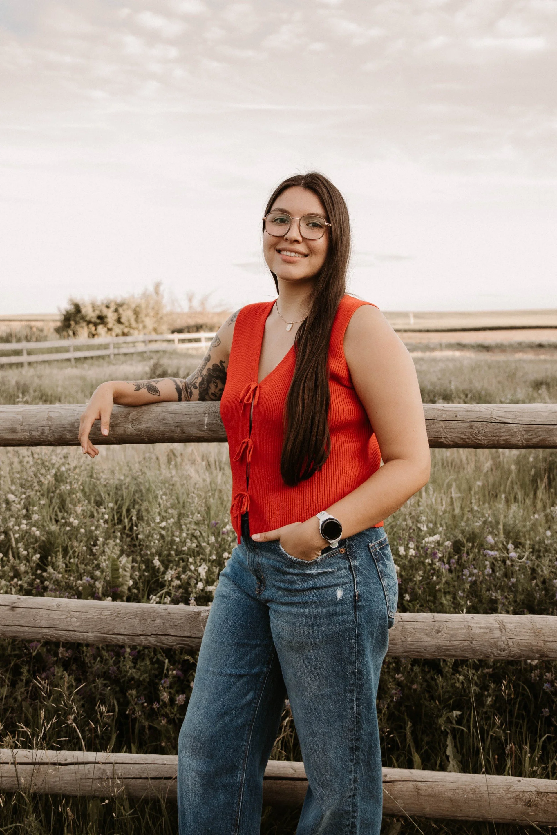 Long brown hair woman wearing a red shirt and jeans leaning on a wood fence smiling
