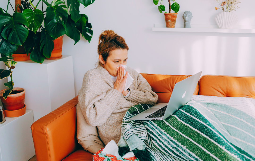 before during and after the flu - woman sitting on the couch working blowing her nose covered in a blanket