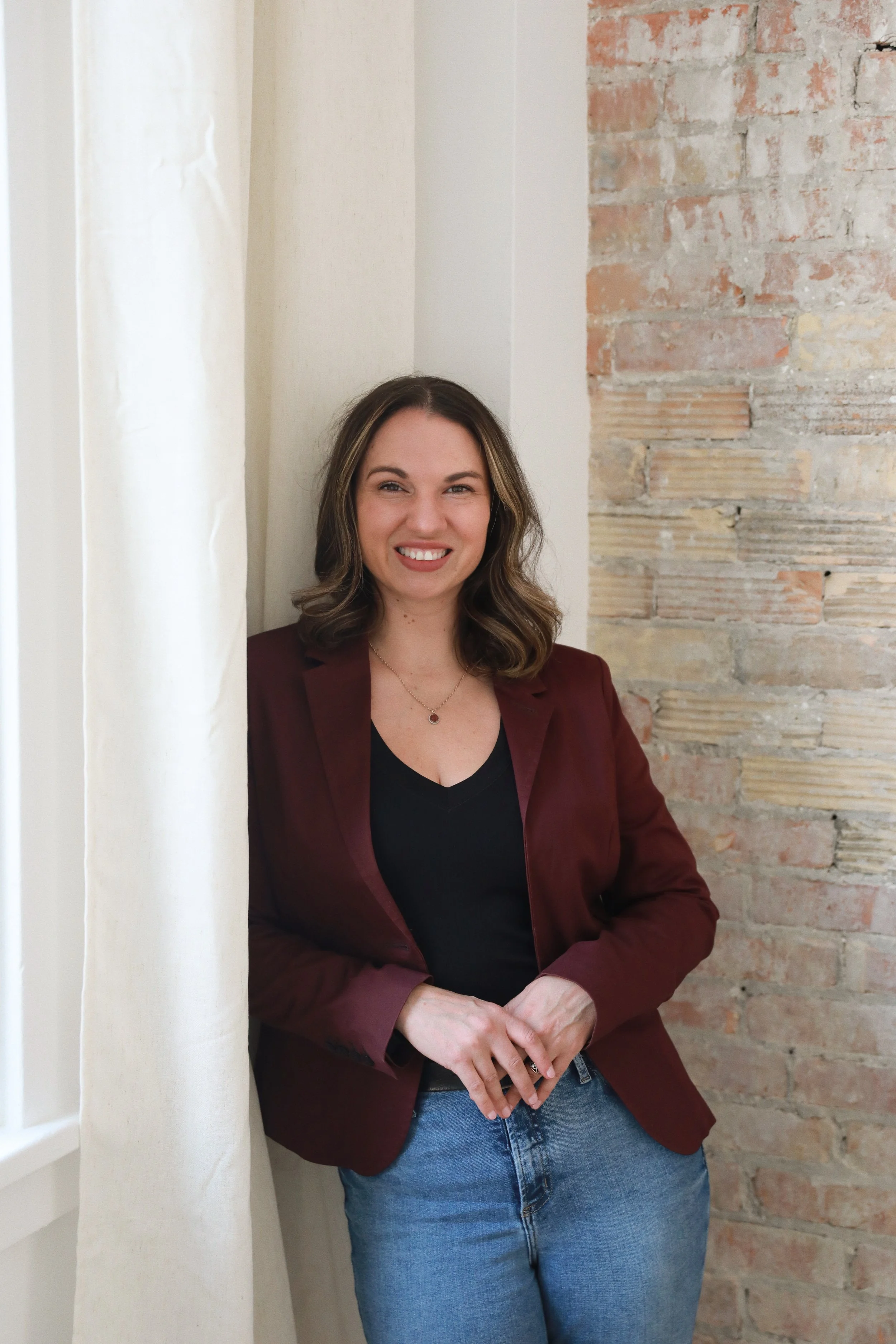a smiling woman with shoulder-length brown hair, wearing a burgundy blazer over a black top and blue jeans, standing in front of a rustic brick wall.