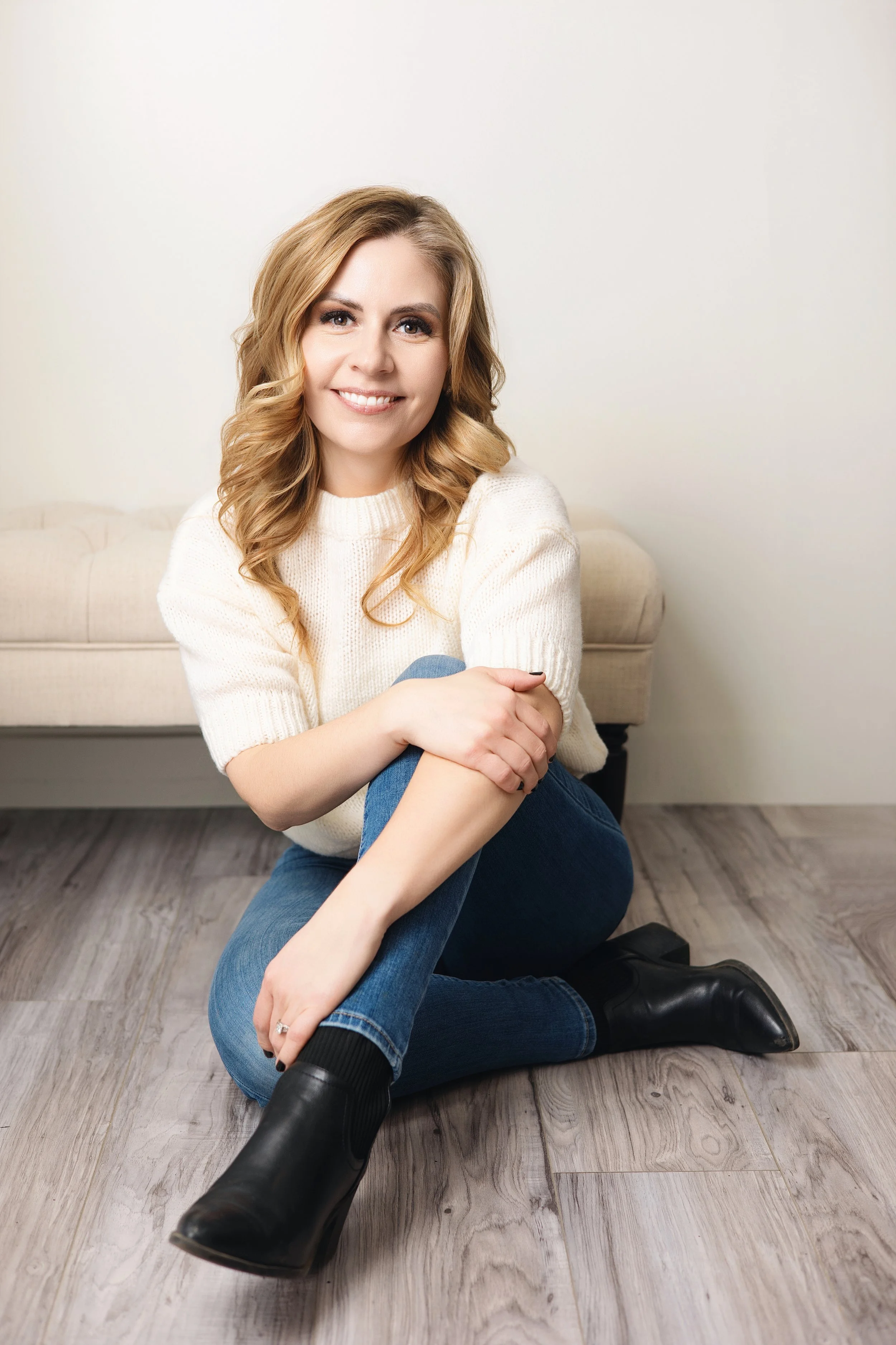 Woman with blonde hair and bangs, wearing a dark top and green pants, sitting on a stool, smiling.