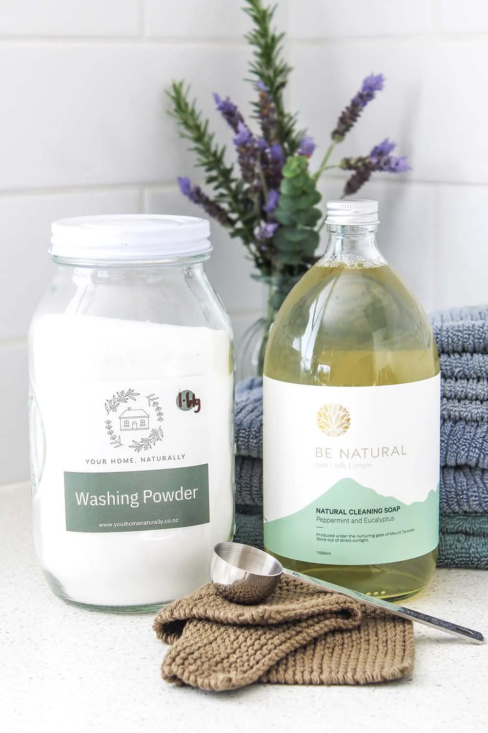 A clear jar of washing powder, a bottle of natural cleaning soap with eucalyptus and peppermint, a measuring scoop on a knitted cloth, and a stack of blue towels with a sprig of lavender in the background.