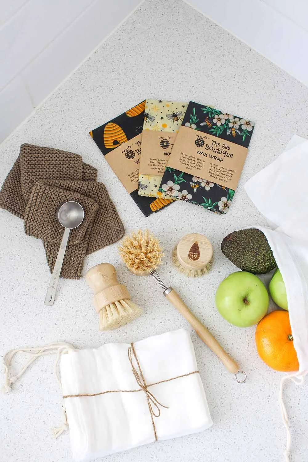 Natural cleaning products, wooden brushes, fresh green apples, orange, avocado, sustainable fabric bags, and beeswax wrap on a white textured surface.