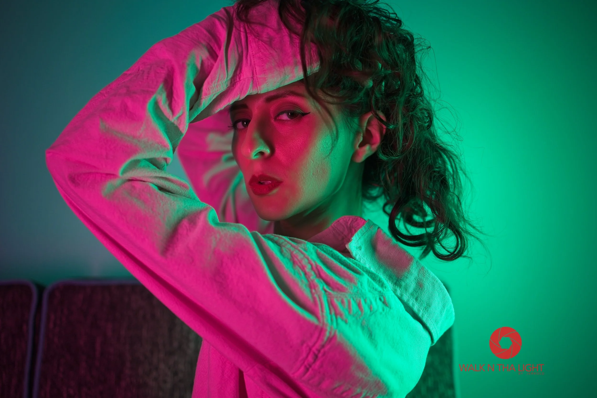 A woman with curly hair wearing a denim jacket is illuminated by pink and green lighting, striking a pose with one hand on her head and looking at the camera.