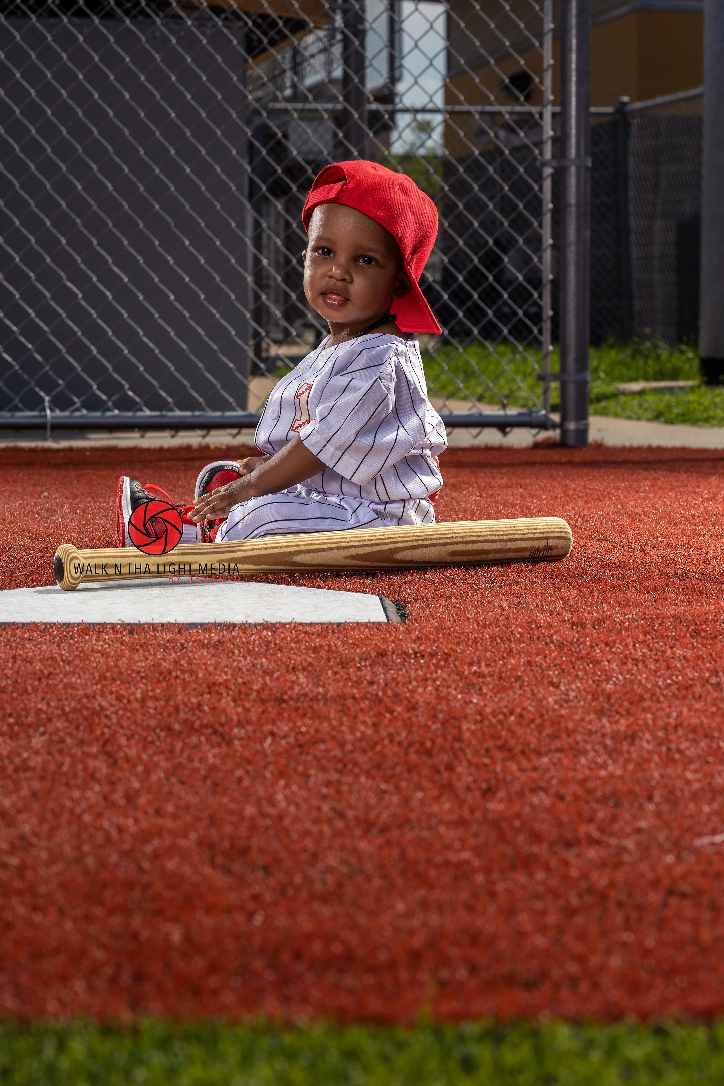 Young child dressed in a baseball uniform with a red cap, sitting on a baseball bat on an outdoor sports field.