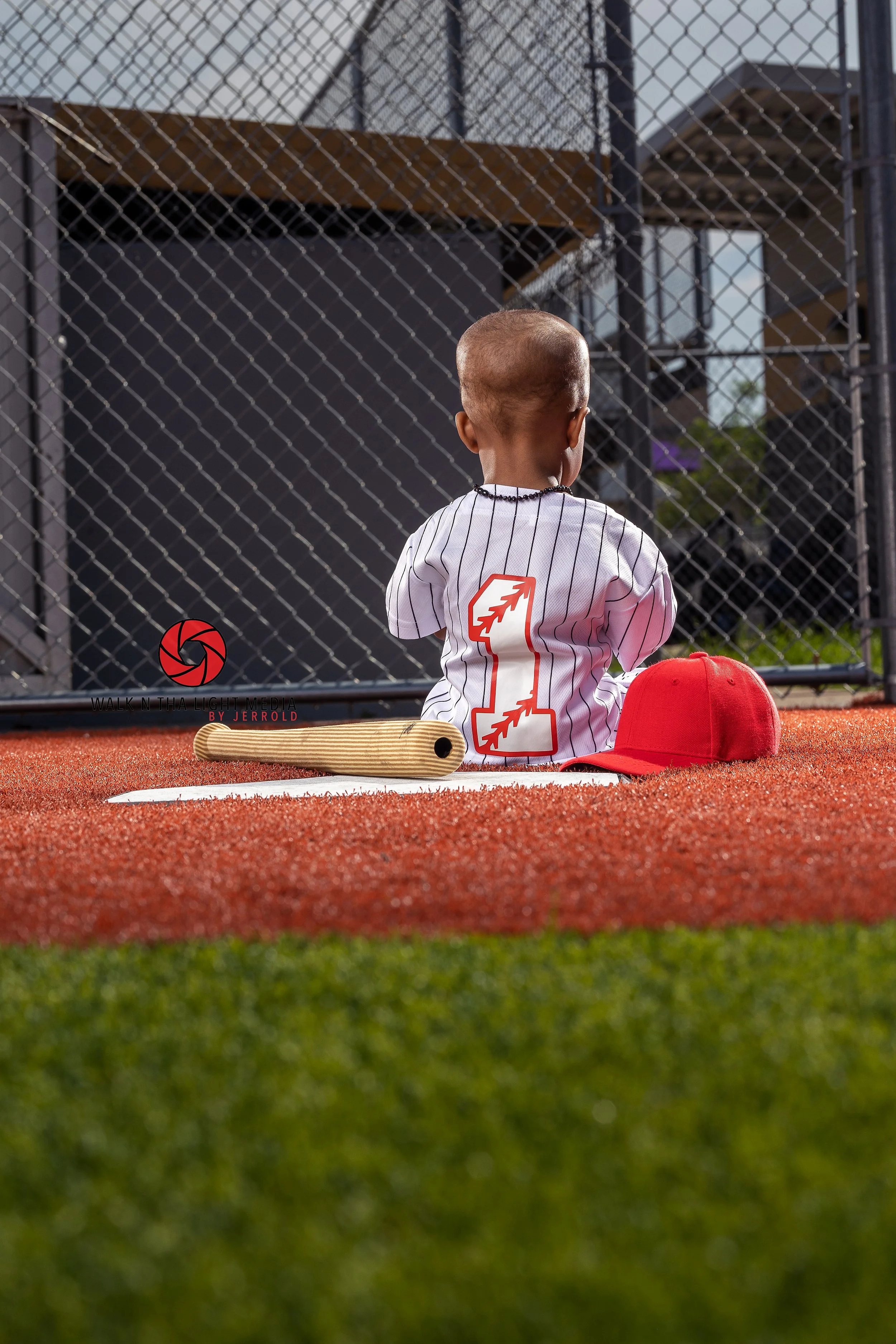 A young boy sits on a baseball field near home plate, wearing a white baseball jersey with a red number 1 on the back, a red cap, and a necklace, with a bat and white glove on the ground beside him, facing a chain-link fence.