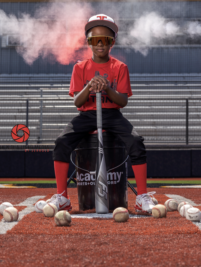A young baseball player in a red shirt, sunglasses, and a cap sitting on a box with a bat, surrounded by multiple baseballs on a baseball field during sunset or practice, with smoke in the background.