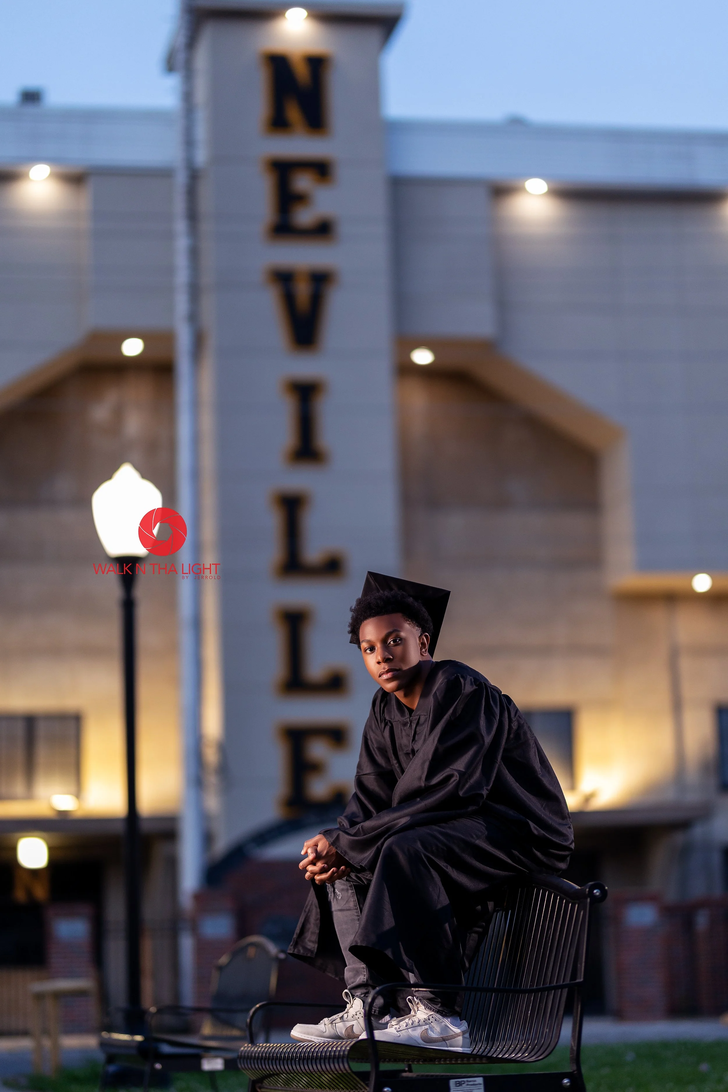 A young graduate in a black cap and gown sitting on a park bench outside a building with a tall sign that reads "Neville" during dusk.
