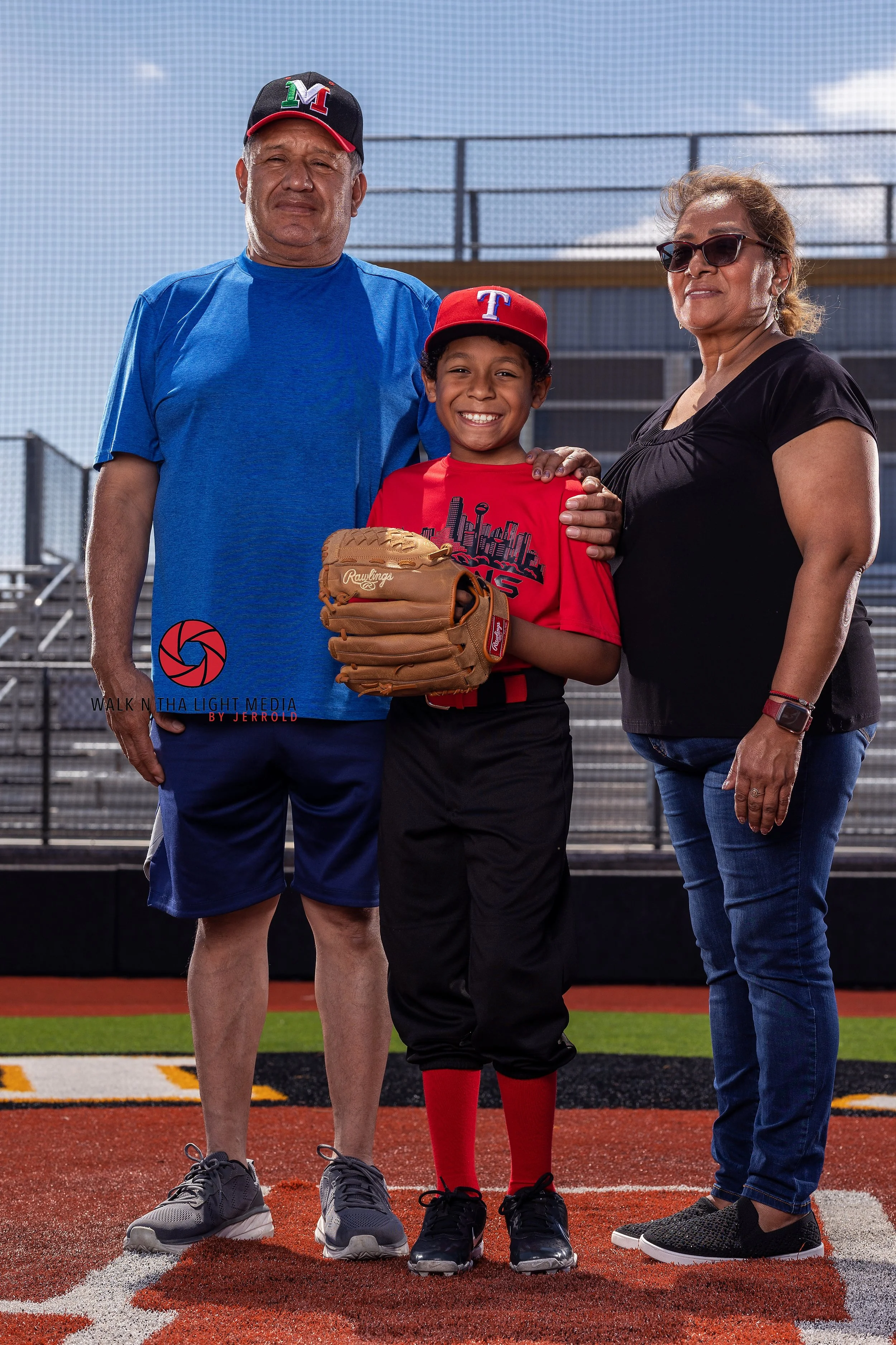 A young baseball player in a red uniform holding a baseball glove, standing between an older man and woman on a baseball field, all smiling at the camera.