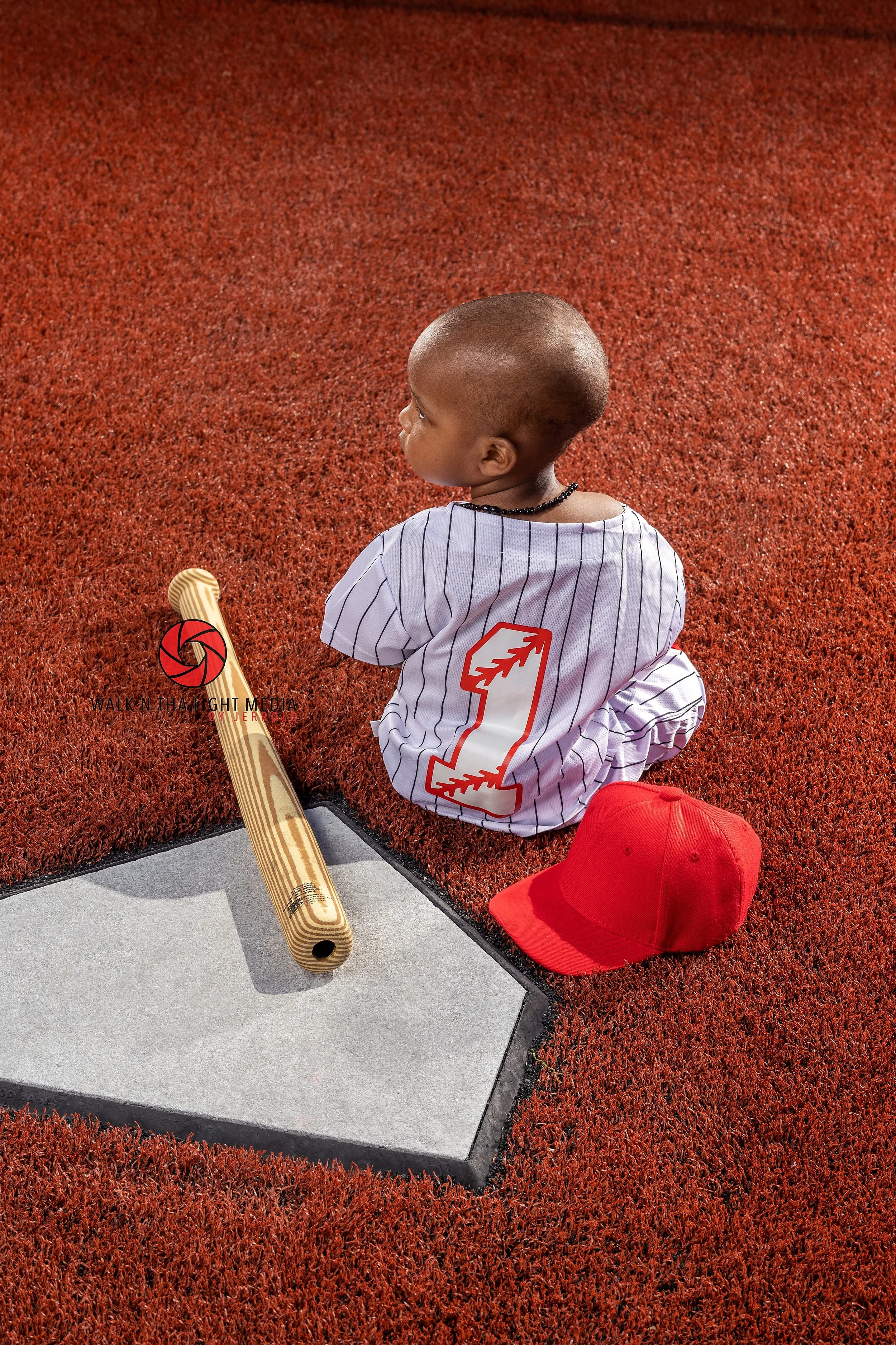 A young boy wearing a baseball jersey with the number 1 on the back, sitting on a red athletic field next to a wooden baseball bat and a red baseball cap.