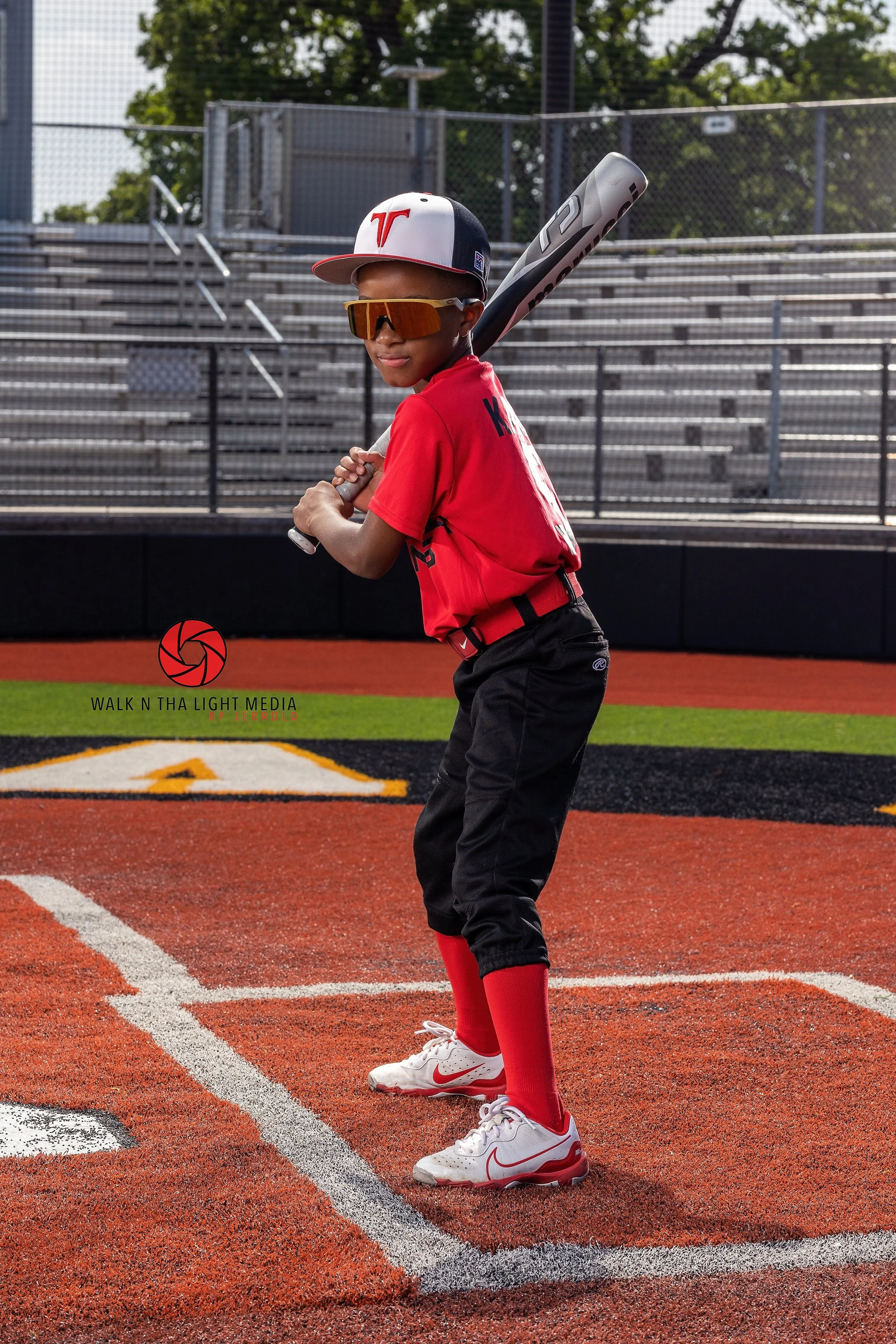 Young baseball player in a red jersey and black pants, holding a bat on a baseball field, wearing a white cap with a red logo, sunglasses, and red socks.