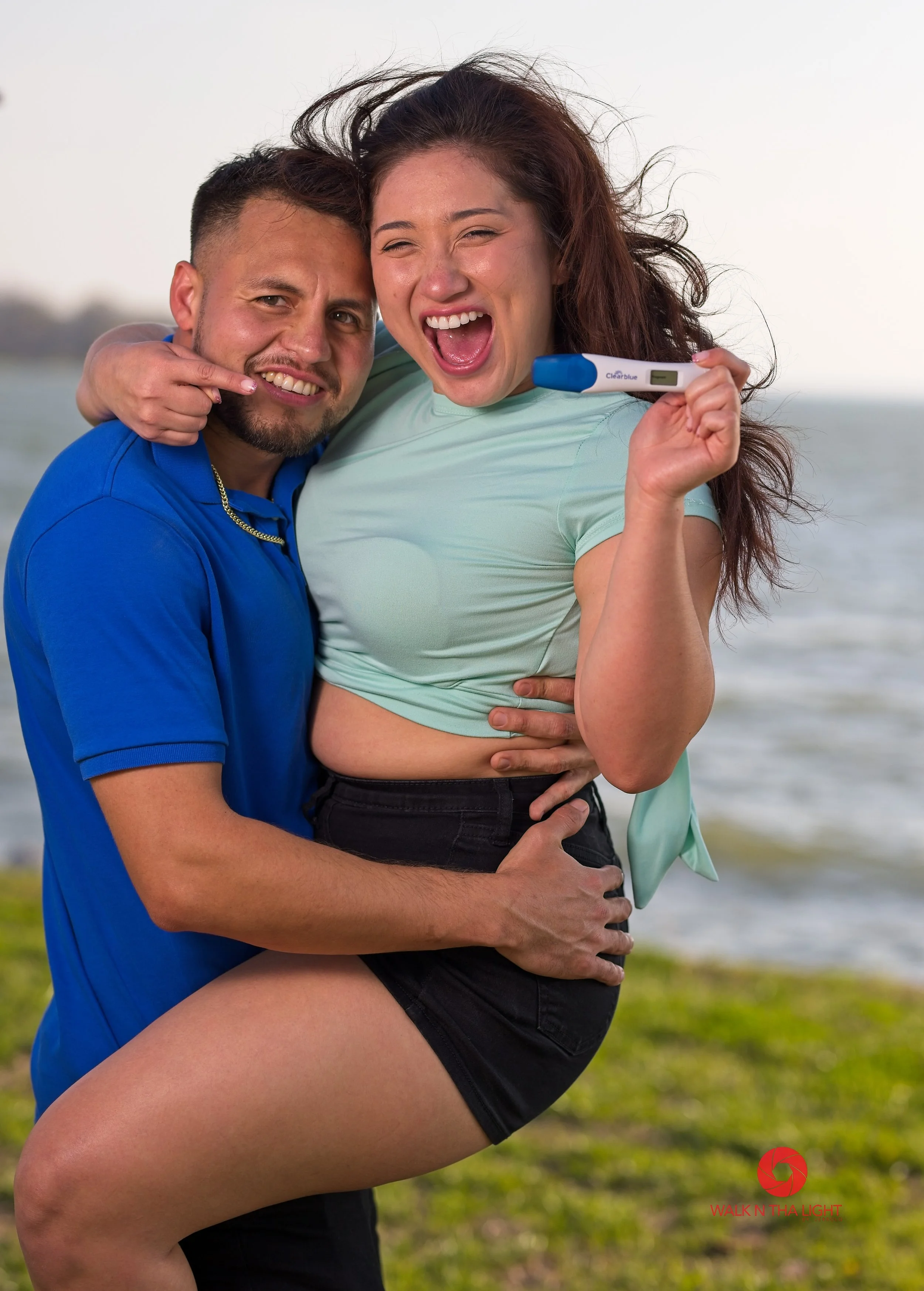 A man and woman outdoors near water, celebrating their pregnancy with a positive pregnancy test, the woman holding it up, both smiling joyfully.