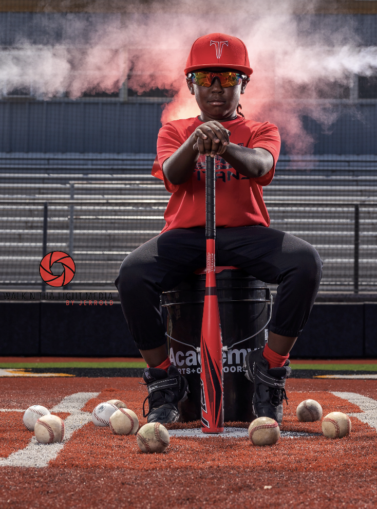 A young baseball player in a red uniform and cap sitting on a bucket on a baseball field, holding a bat, surrounded by multiple baseballs, with a smoky background.