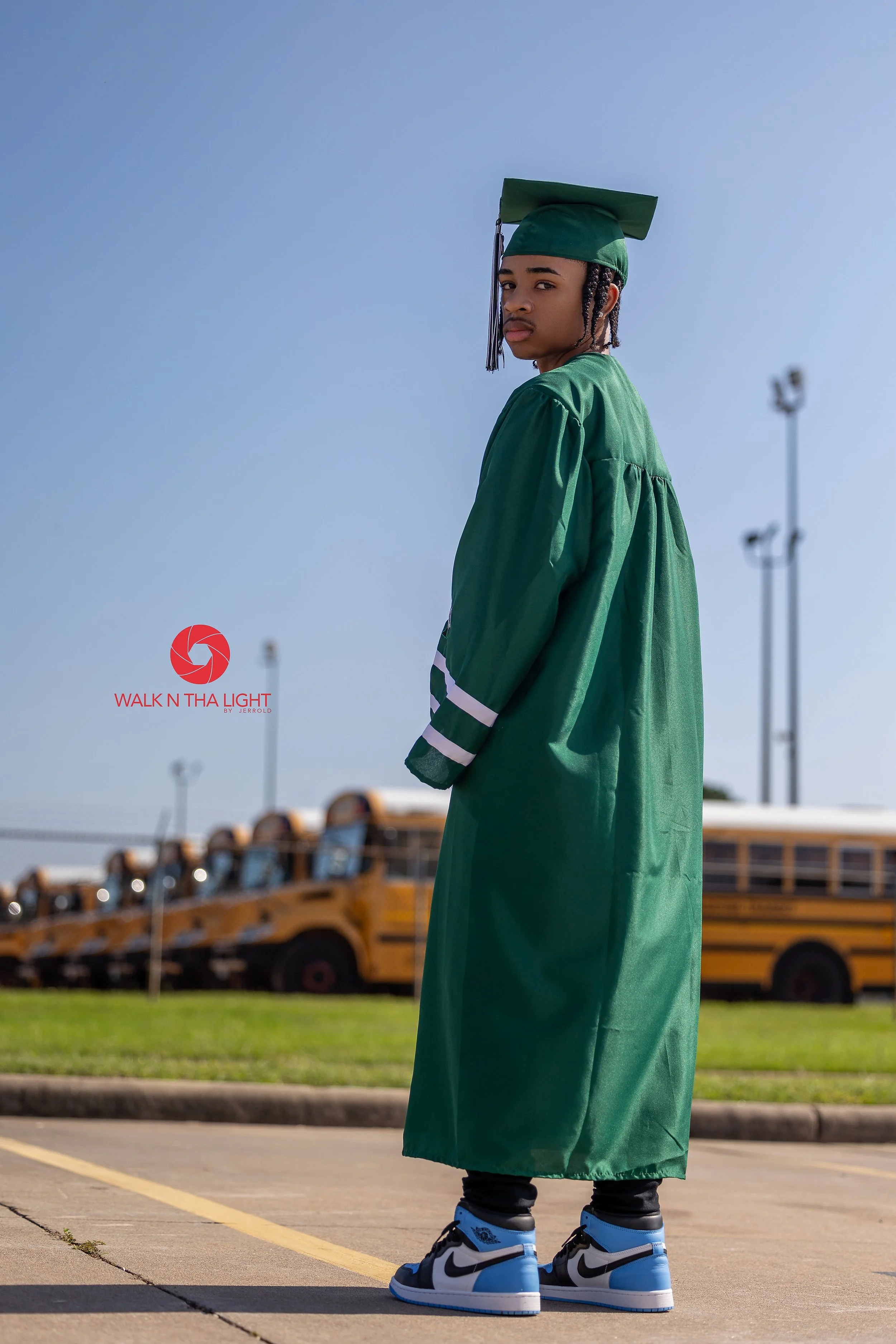 A young man in a green graduation cap and gown standing outdoors near yellow school buses, with a clear blue sky in the background.