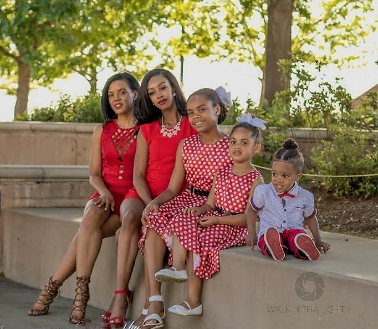 Family sitting outdoors on a bench near trees, dressed in red and polka dot dresses, with two young girls wearing bows in their hair and a woman with long hair in a red dress, all posing for a photo.