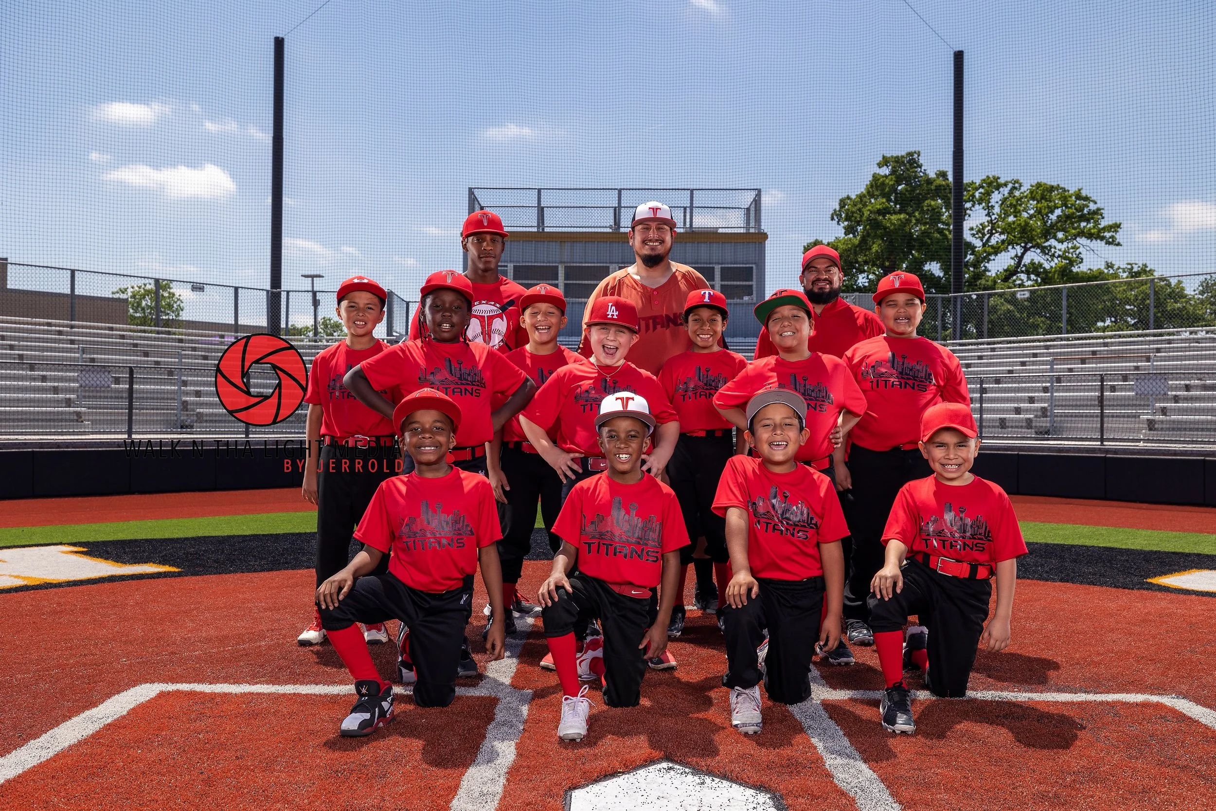 Youth baseball team wearing red jerseys and caps posing on the field with coaches, baseball diamond in the background, during daytime.
