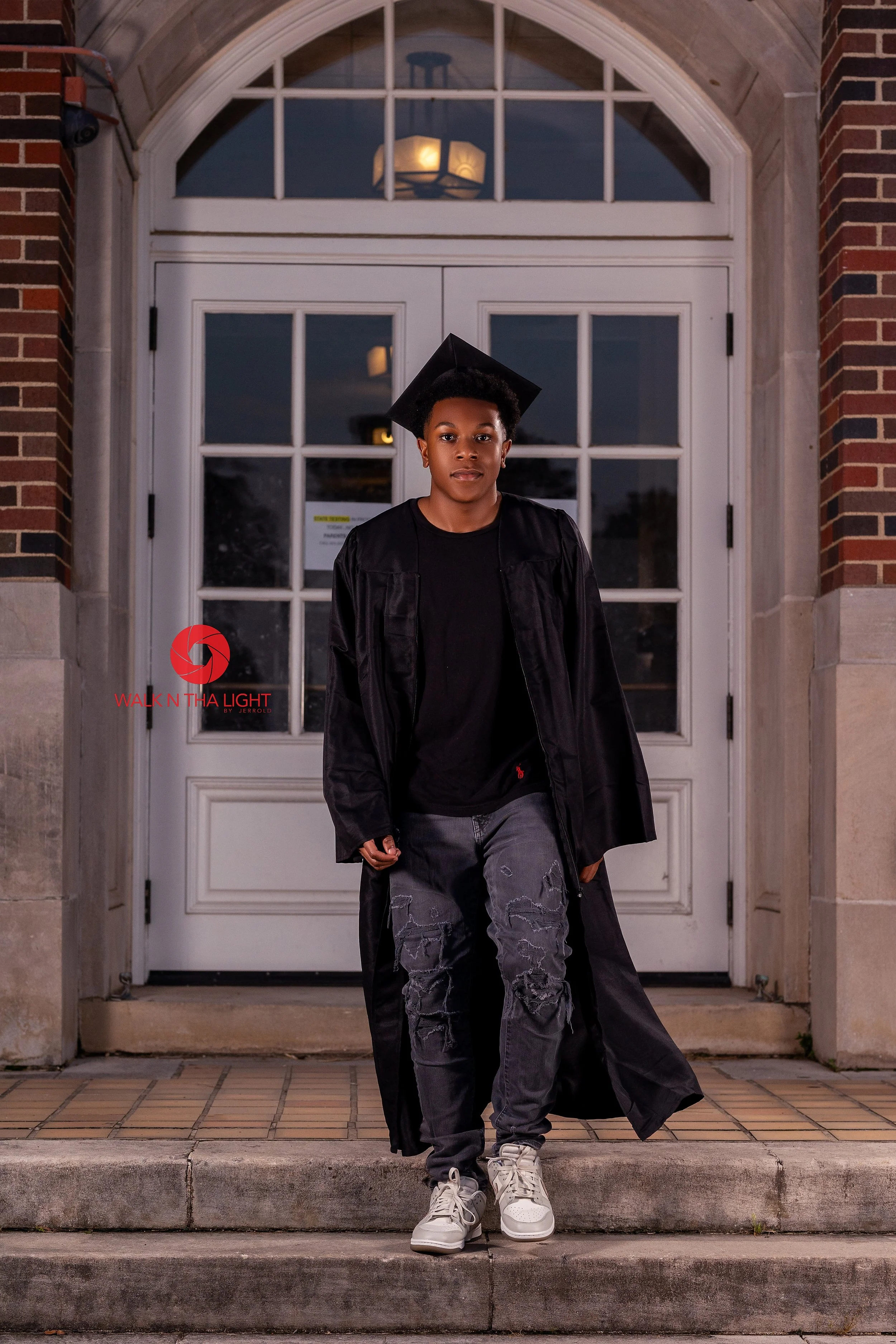 Young person in graduation gown and cap standing on steps in front of a white door with brick and stone framing, during dusk or evening.