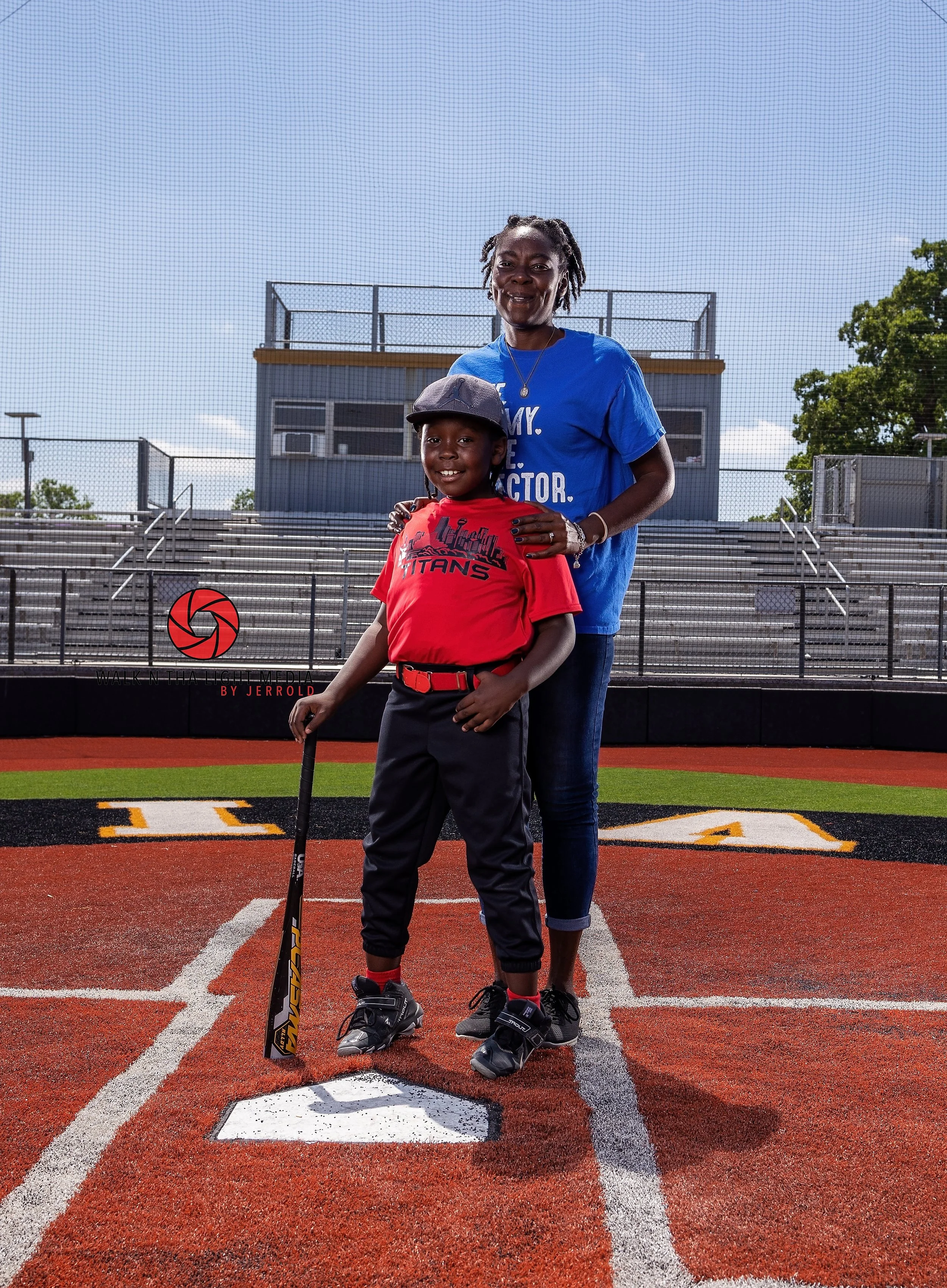 A woman and a young boy smiling on a baseball field. The boy is holding a baseball bat and wearing a baseball uniform. The woman is standing behind him, with her hand on his shoulder, and they are in the middle of a sports stadium with empty bleacher
