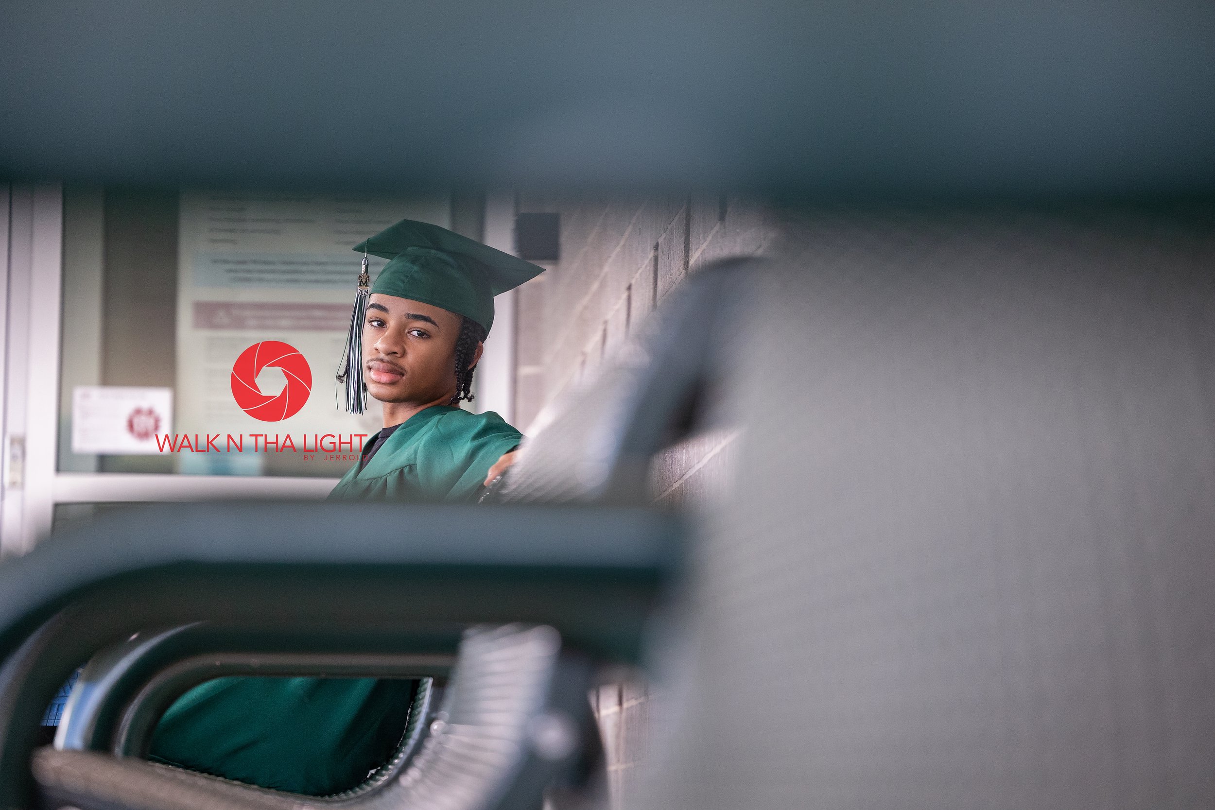 Young man in a green graduation cap and gown sitting on a bench at a school or college campus.