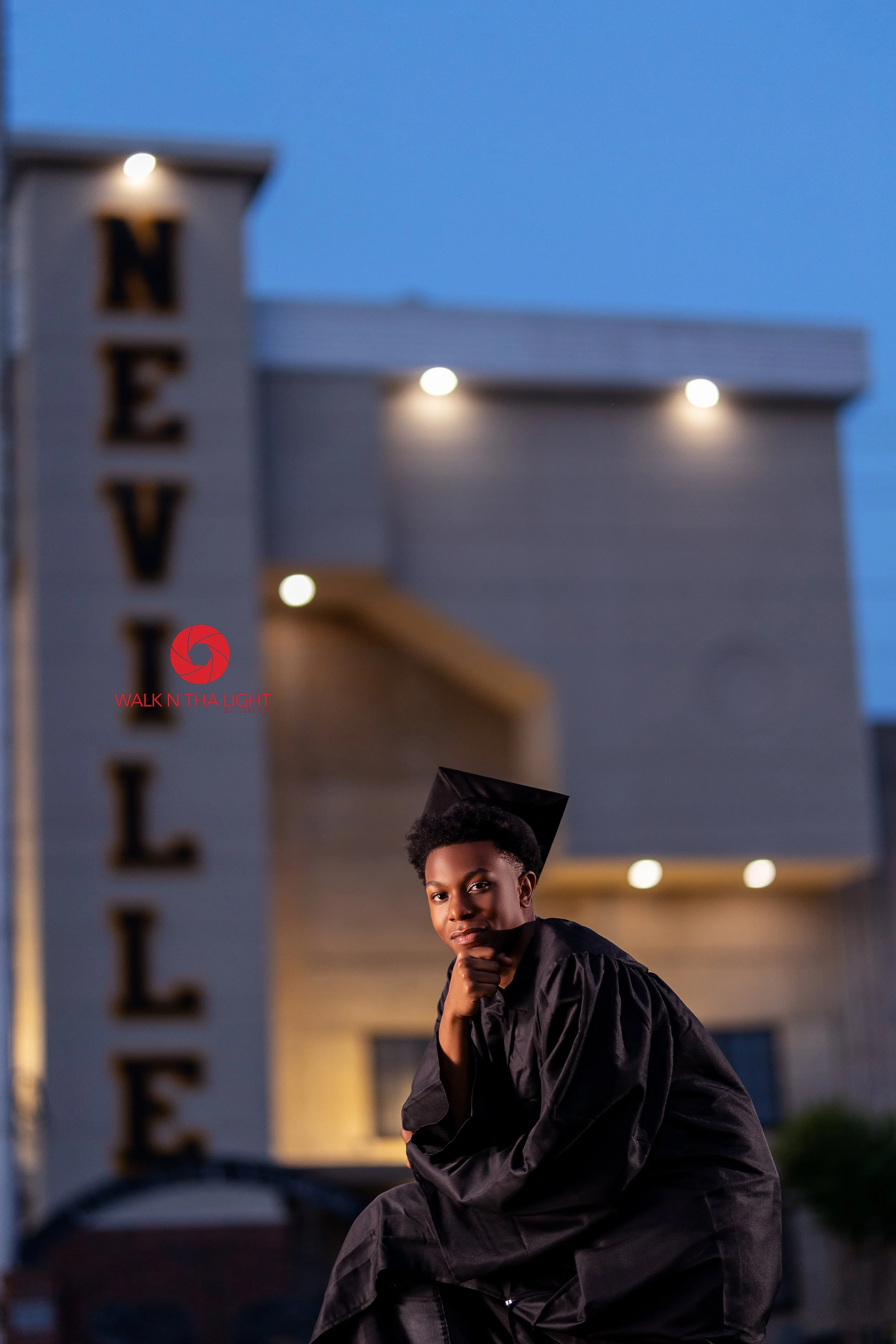 A young man in a graduation cap and gown sitting outdoors at dusk with her chin resting on her hand in front of a building with a sign that reads "NEVELL" illuminated at night.