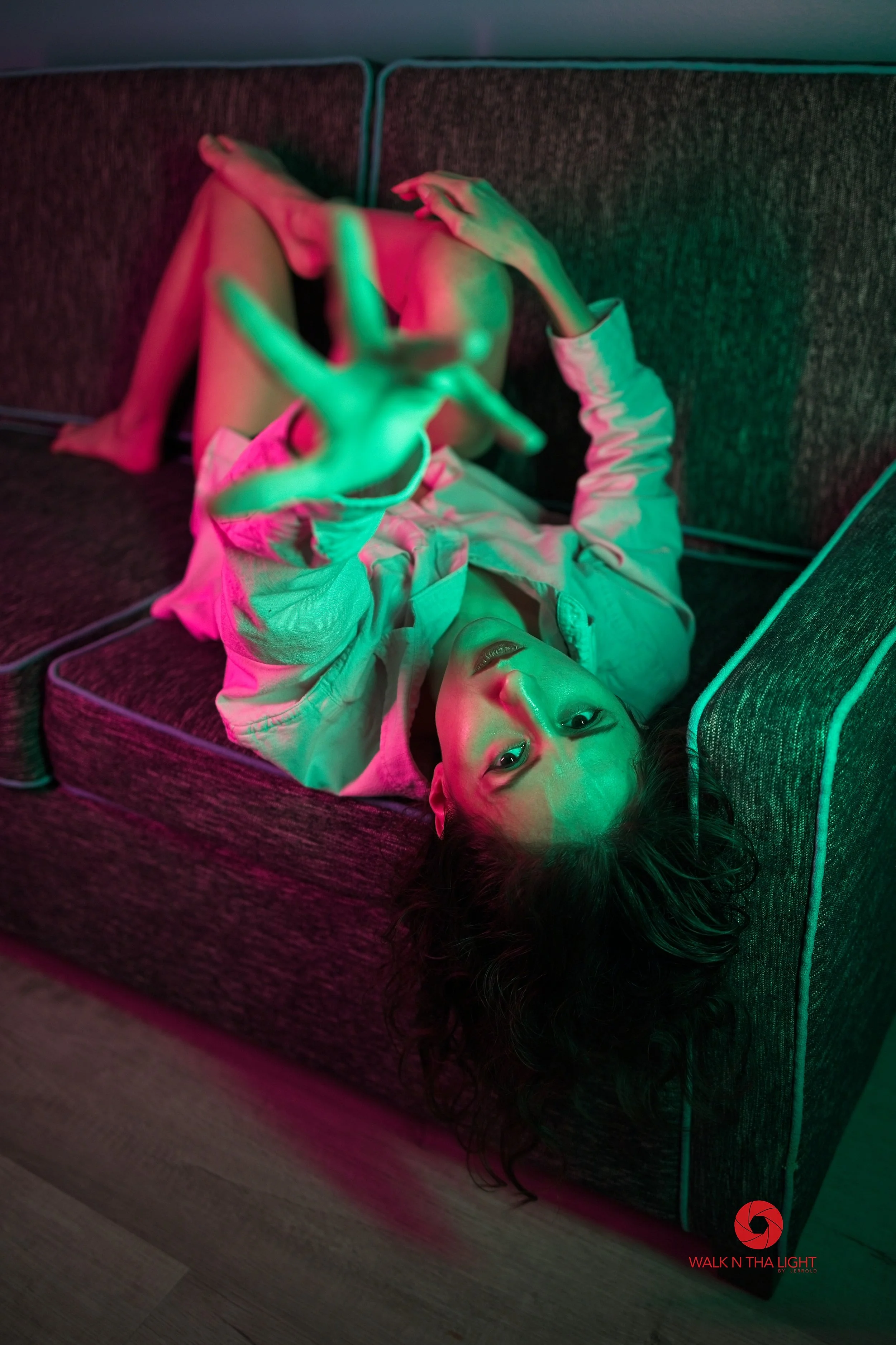 A young woman with long, dark, curly hair lies on a dark upholstered couch with green and pink lighting. She is looking up at the camera, smiling slightly, with one hand extended toward the lens and the other resting on the couch. She is wearing a li
