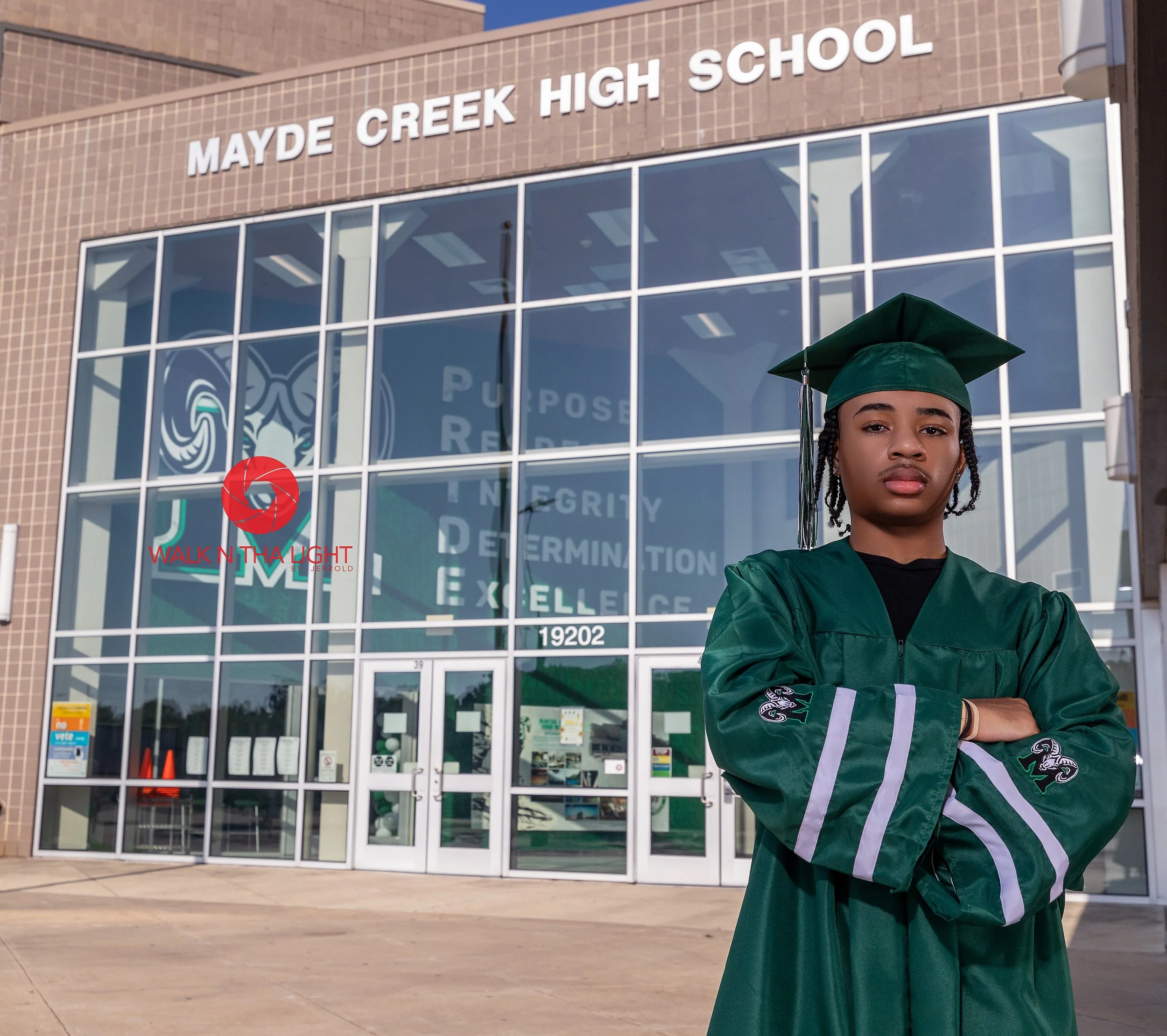 A young man in a green graduation gown and cap stands with arms crossed in front of Mayde Creek High School building, with large glass windows and the school's emblem. The school name is visible at the top of the building.