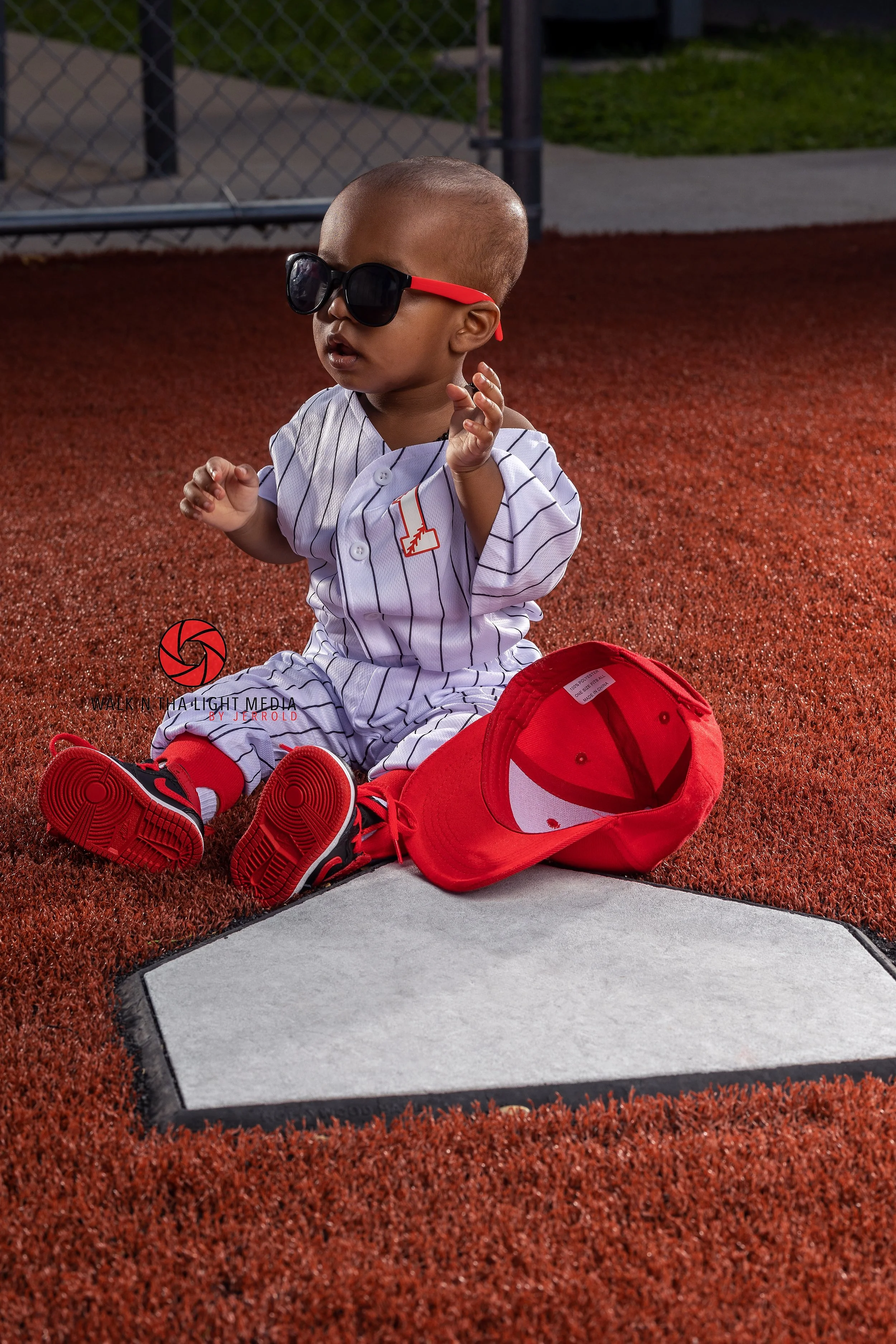 Young child sitting on a baseball field wearing sunglasses, a baseball uniform, red and black sneakers, with a red cap lying on the ground beside them.