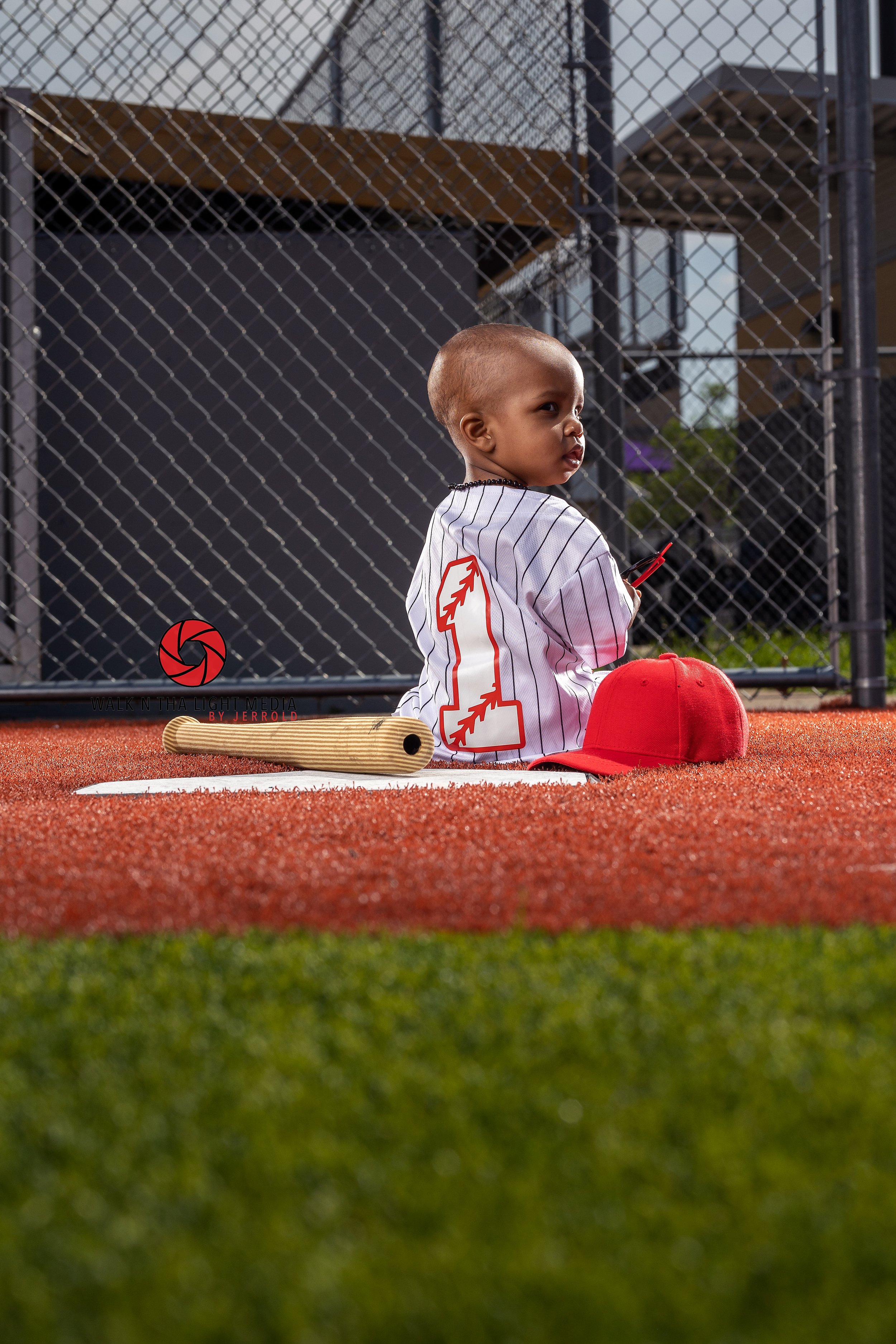 A young boy sitting on a baseball field, wearing a striped jersey with a large red and white logo on the back. A red baseball cap and a wooden bat are placed on the ground nearby. He is holding a small red object and appears to be looking over his sh