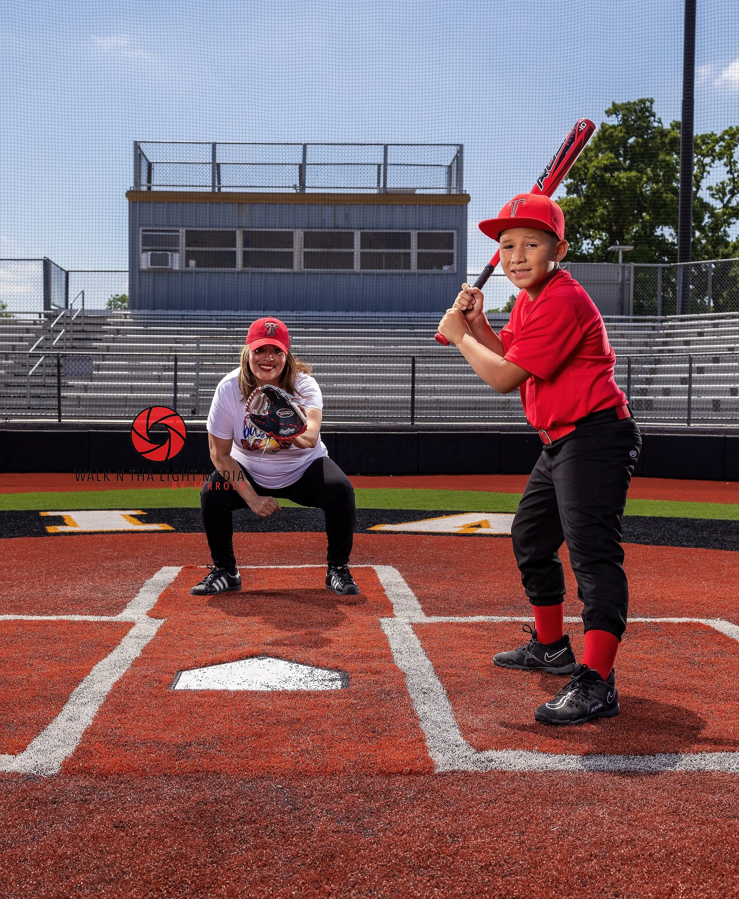 A young boy in a red baseball uniform and cap holding a bat, standing at home plate on a baseball field, facing an adult woman catcher crouching behind home plate with a glove, both smiling. The background features empty bleachers and greenery.