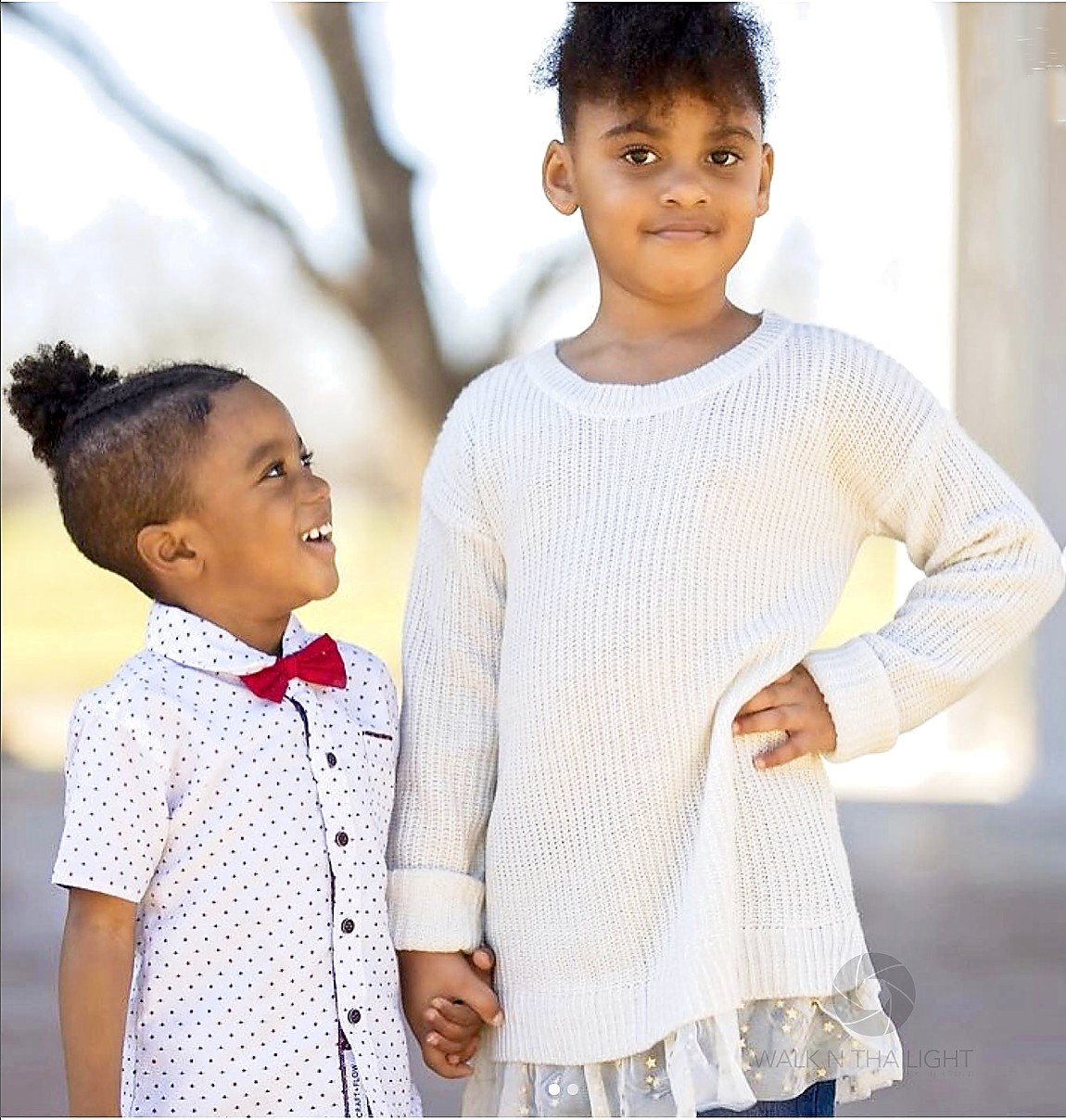 Two young Children, one taller and older, holding hands outdoors, smiling, with a blurred background of trees and sky.