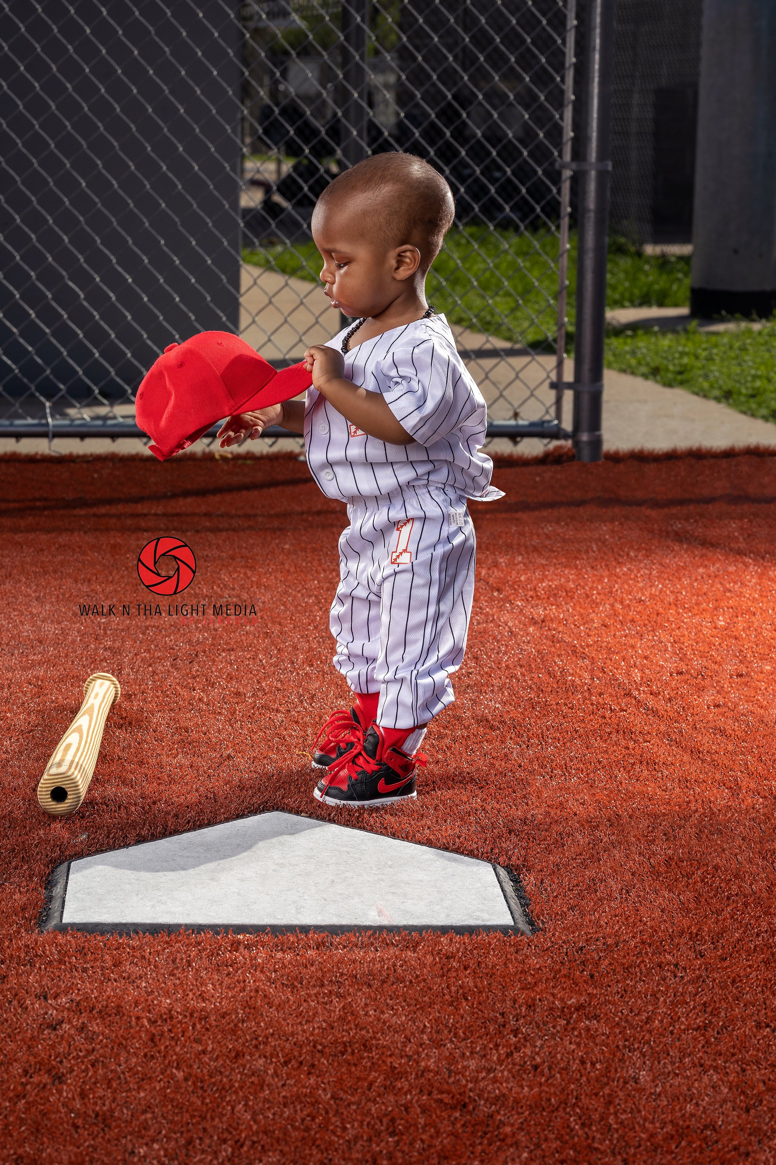 A young boy dressed in a baseball uniform holds a red baseball cap while standing on a baseball field near home plate, ready to bat.
