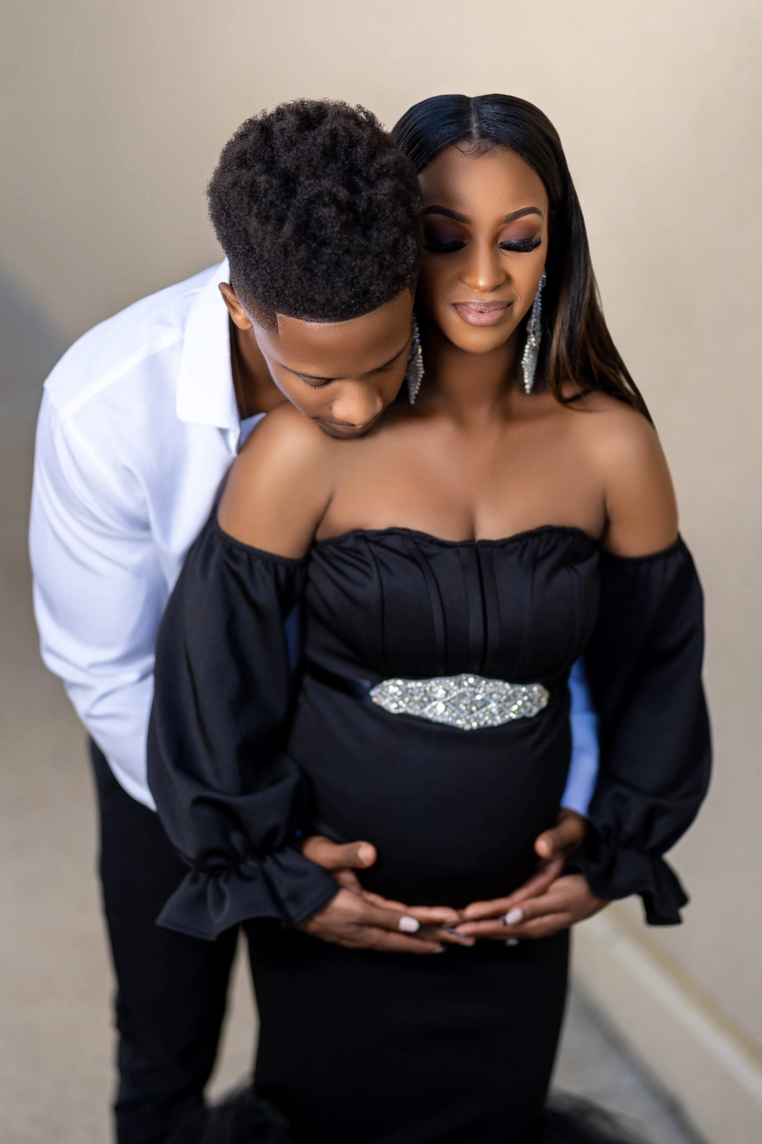 A couple, with the man kissing the woman's shoulder, standing close with hands on her pregnant belly, in an intimate embrace.