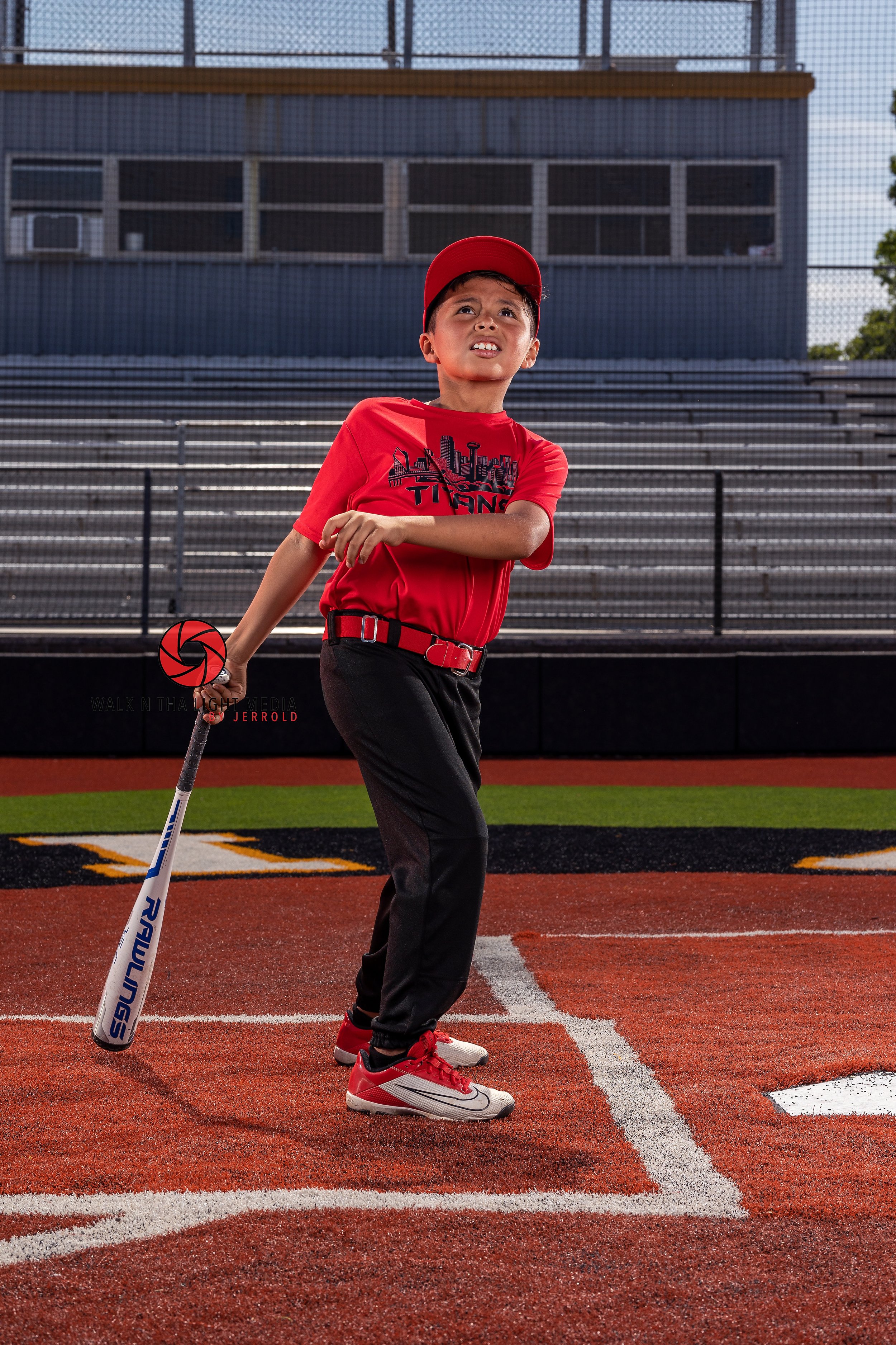 A young boy in a red baseball uniform holding a baseball bat on a baseball field, with metal bleachers and a building in the background.