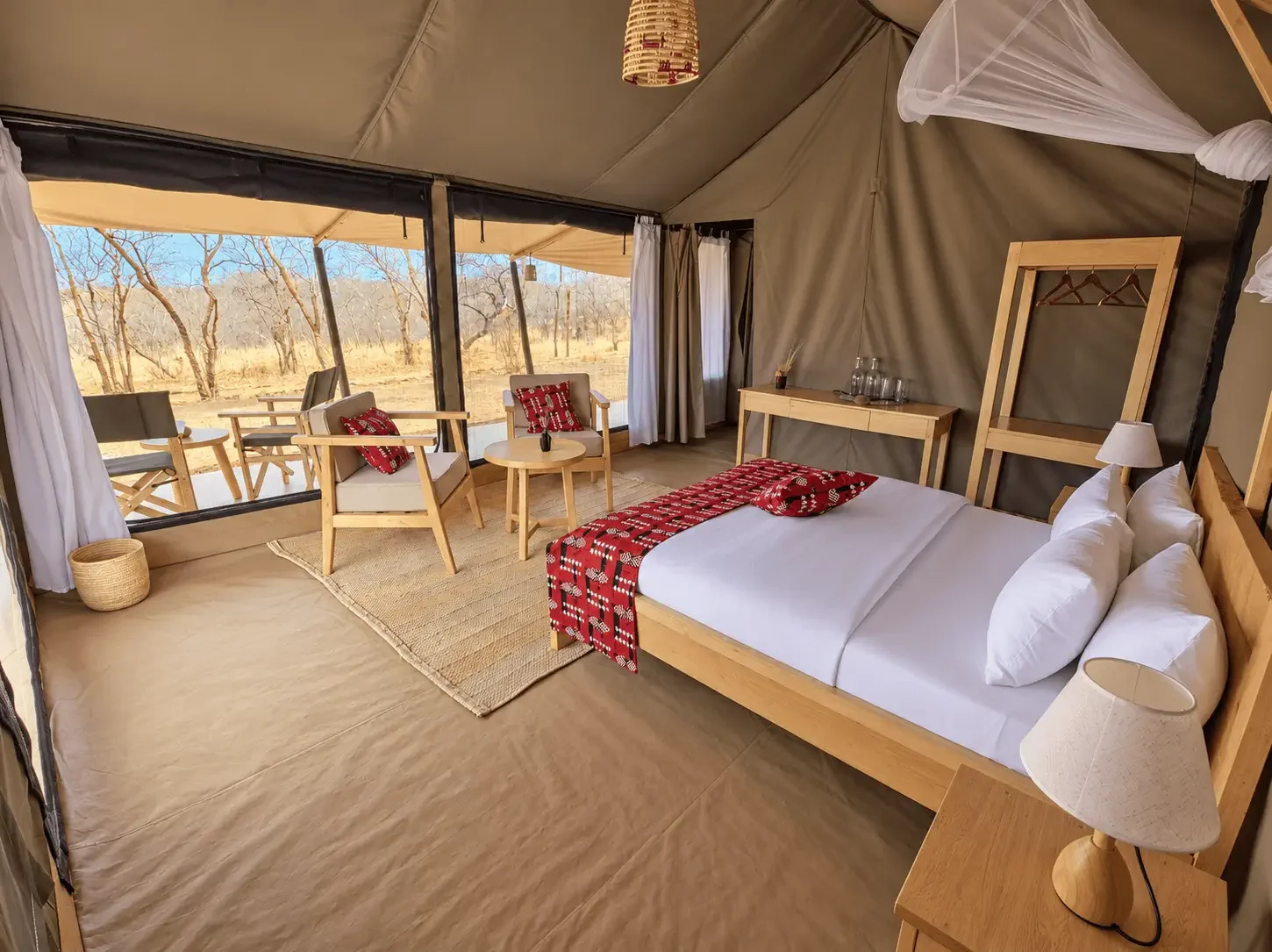 Ein komfortables Schlafzimmer in einer Safari-Lodge mit einem großen Bett, einem kleinen Tisch, Sesseln, einem Schrank, einem Teppich und einer großen Fensterfront mit Blick auf die afrikanische Savanne.