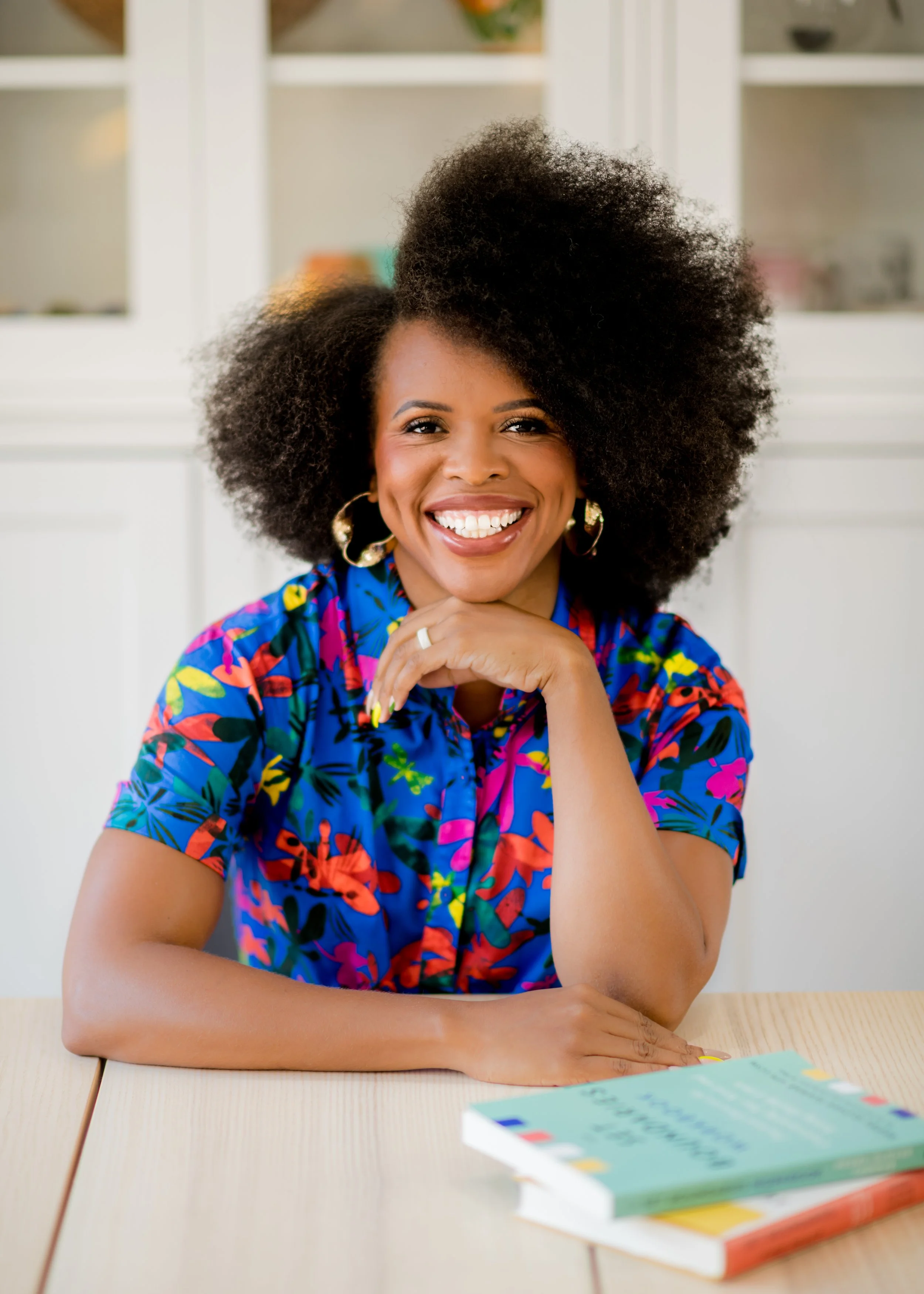 Nedra Tawwab is sitting at a desk smiling in a bright and colorful shirt. She is sitting next to two of her books, Set Boundaries, Find Peace and The Set Boundaries Workbook.