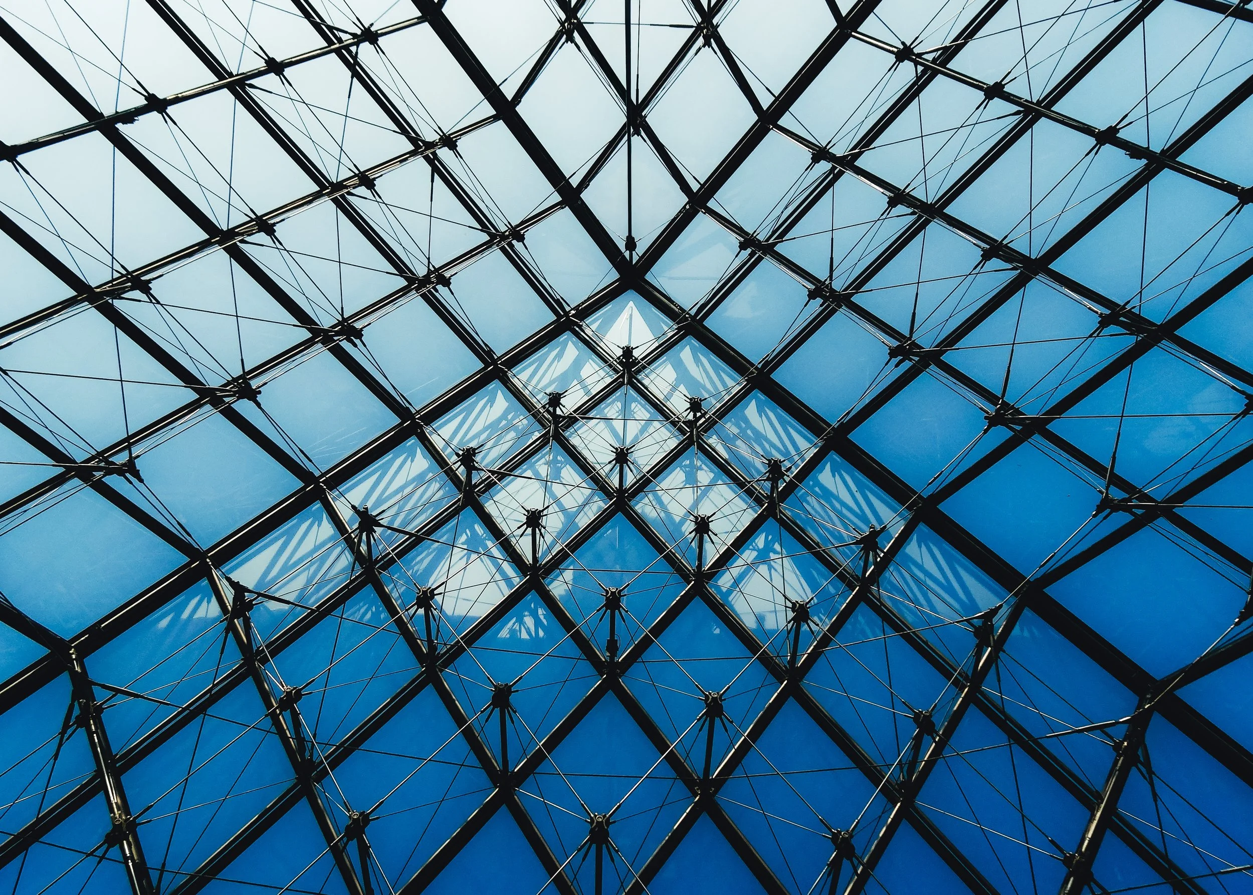 Looking up at a glass ceiling with a geometric steel framework and blue sky visible through the glass panels.