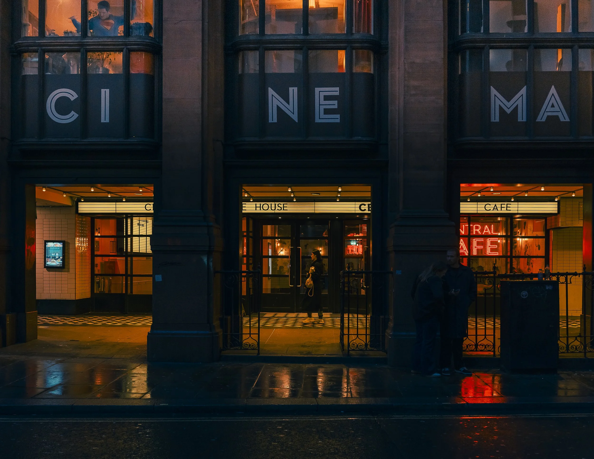 Nighttime view of a building with large windows spelling out "CINEMA" and a neon sign that says "CINEMA CAFE" inside. People are standing outside and inside, with reflections on the wet sidewalk.