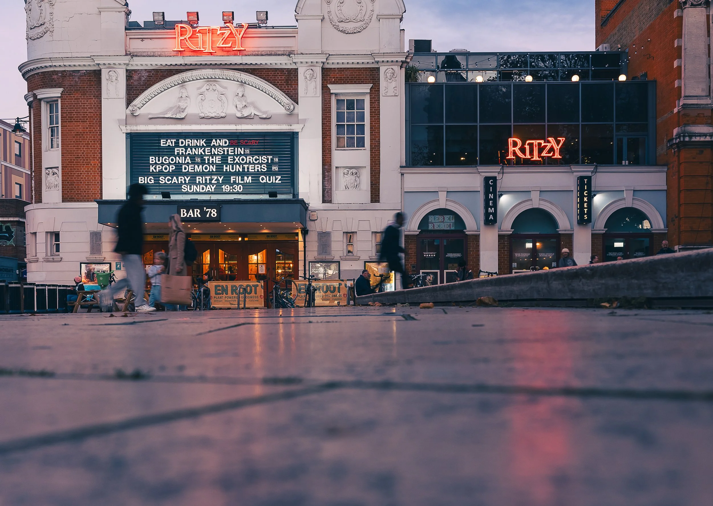 This is a street view of the Regal Cinema building with a red neon sign that says 'RITZ' on top. The building has a classic design with a marquee announcing events, including a film quiz and horror movies. There are people walking and sitting in fron